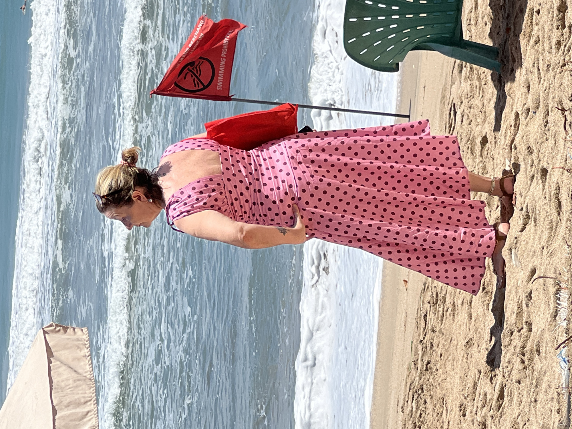In Kuta, Indonesia, a woman stands on a sandy beach near the ocean, wearing a pink polka dot dress and holding a red bag. The ocean has waves and a red flag is planted in the sand. A green plastic chair is to the right of the woman.