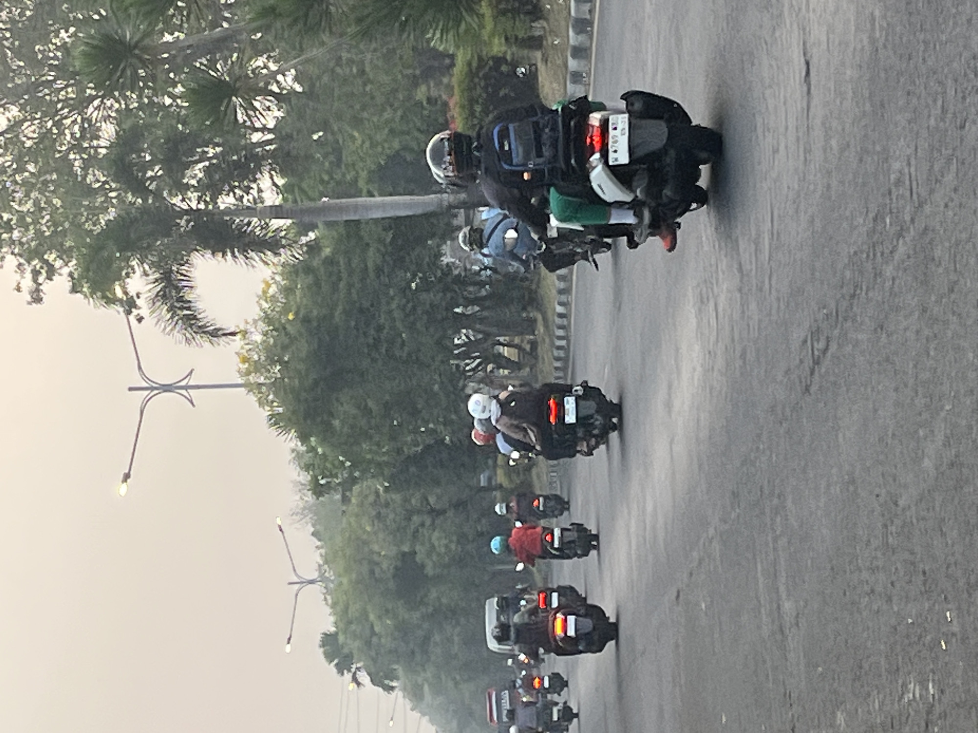 The image captures a street scene in Kludan, Indonesia, likely during the day with overcast conditions. A group of people are riding motorcycles on a paved road. The motorcycles are a mix of models, with several appearing to be scooters. Most riders are wearing helmets, and some are carrying backpacks. The road has lane markings, and on the sides, there are trees and palm trees, suggesting a suburban or semi-rural setting within the city. Lampposts are visible in the background. The overall atmosphere is one of everyday traffic and transportation.