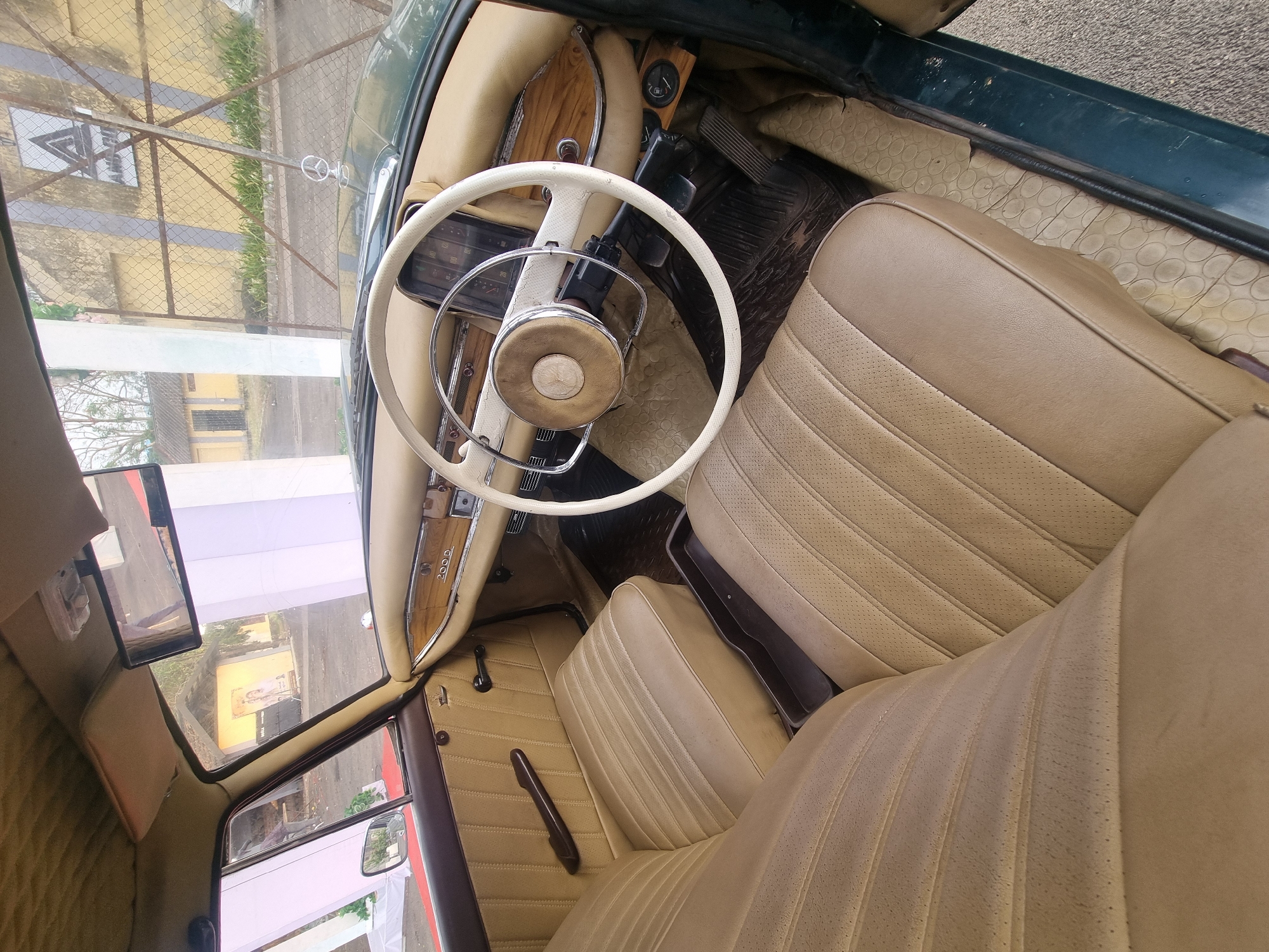 This media file showcases the well-preserved interior of a vintage Mercedes-Benz car, identified by the "200 D" badge on the passenger side dashboard and the Mercedes-Benz logo on the steering wheel and as a hood ornament. The car features a cream or tan-colored interior, with perforated leather upholstery on the seats, detailed with vertical stitching. The dashboard is adorned with polished wood trim, and the steering wheel is white with a chrome horn ring.
Through the windshield, a daytime scene is visible, including a chain-link fence, a yellow building, and greenery, suggesting the car is parked outdoors in a residential or semi-urban setting, consistent with the location context of Kalamassery, India. A white triangular sign is also discernible in the background. The driver's footwell contains black rubber floor mats over a patterned cream flooring, with the accelerator, brake, and clutch pedals clearly visible. An auxiliary gauge is mounted near the driver's feet area. The passenger door panel also features wood trim and a manual window crank. An additional, smaller mirror is mounted on the windshield frame. The car's exterior, partially seen through the windows and door sills, appears to be a dark green color. No people are visible, and the car appears stationary.
