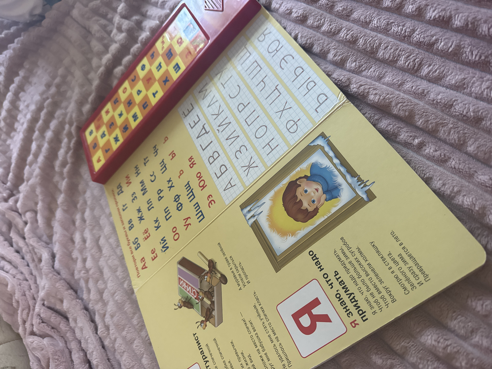 An open book is laid on a pink plush blanket. The book has a yellow cover and its pages are filled with text and illustrations, likely for children. On the left page, there is an illustration of ants next to a matchbox with the word "СПИЧКИ" (matches) written on it. Below this, there is a large red letter "Я" inside a white square. Further down are lines of text describing the letter "Я" and a poem. On the right page, there is a grid with Russian alphabet letters. Adjacent to the book is a red and yellow electronic device with buttons also displaying letters of the alphabet. The device appears to be a toy for learning the alphabet. The background is a textured pink blanket. There is no indication of the city of Кашино, Russia.