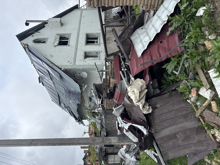 A damaged house with visible bullet holes and broken windows stands amidst debris. Solar panels are affixed to a heavily damaged, partially collapsed roof. Metal sheets and fragments of building materials are scattered across the ground. The devastation is extensive, hinting at a violent event. A utility pole is present in the foreground. Green foliage is visible, contrasting with the destruction.