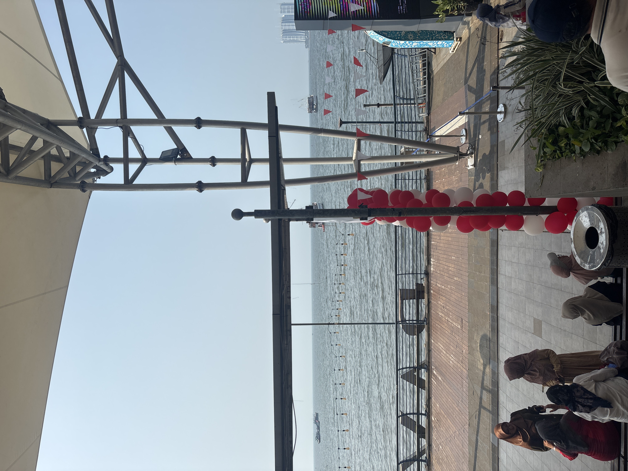 This is a photograph taken at Ancol Beach City, North Jakarta, Indonesia, on a sunny day with a clear sky. In the foreground, a group of women wearing hijabs are gathered, some seated on benches, some standing. They appear to be in conversation. To the left, a string of red and white balloons forms an archway. In the middle ground, a wide expanse of the sea is visible with several boats on the water. A railing runs along the waterfront, and the ground is paved with bricks. In the background, a tall metal structure supporting a white canopy dominates the scene, with buildings visible in the distance. The overall impression is one of a public space by the sea, possibly a promenade or an outdoor venue.
