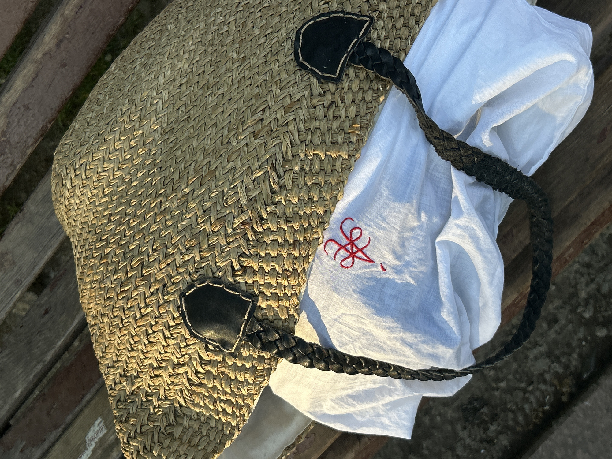 The image shows a woven straw tote bag with black leather handles resting on a weathered wooden surface, possibly a bench. A white cloth with a small red embroidered design is partially tucked inside the bag. The scene appears to be outdoors, judging by the natural light and the adjacent concrete surface.