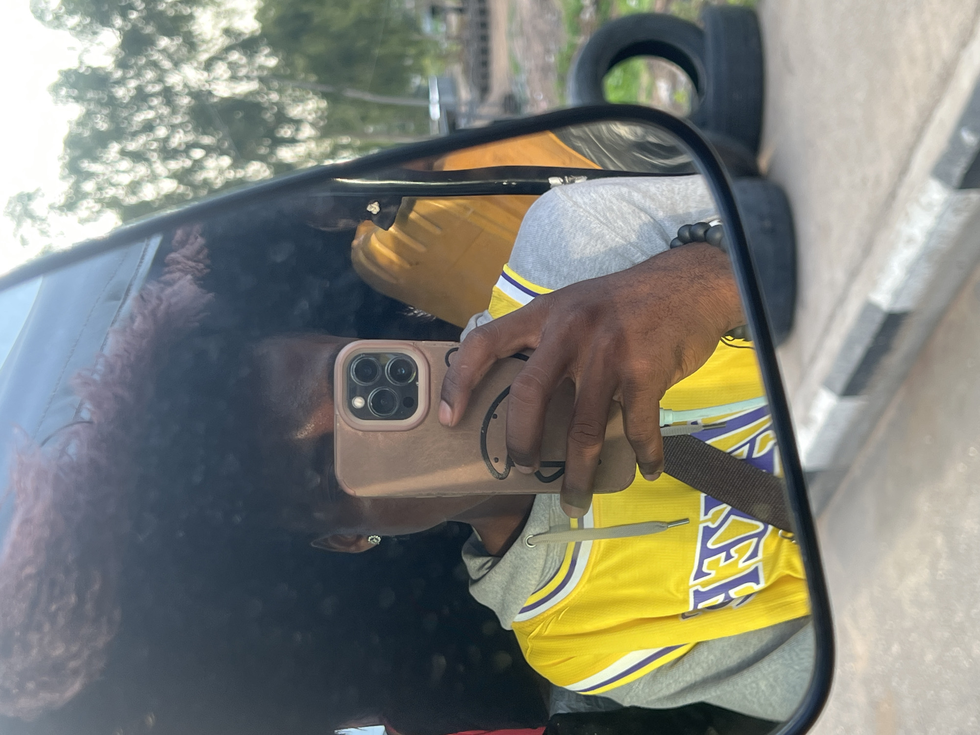 The image is a selfie taken in a side mirror, showing a reflection of a person wearing a yellow Lakers jersey over a grey hoodie. The person's dark hair is visible in the reflection. The person is holding a tan-colored smartphone with multiple camera lenses. A seatbelt is visible across the jersey. In the background, visible through the mirror, are tires stacked on the side of the road. The location is suggested to be in the city of Kporva, Nigeria.