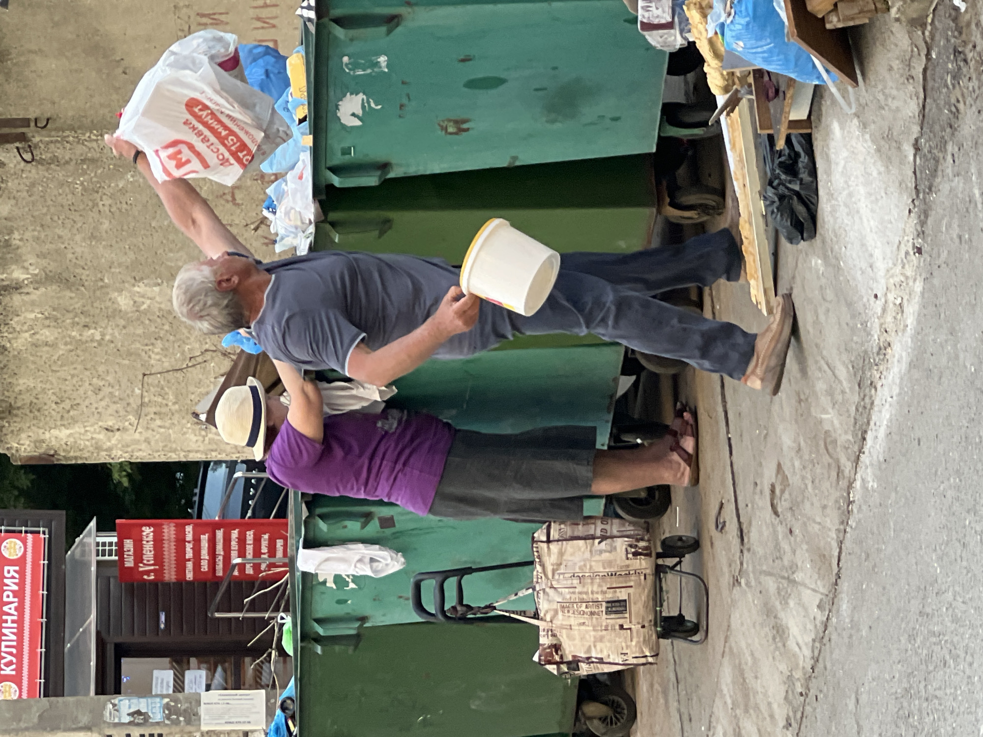 In Krasnodar, Russia, a man and a woman are shown near several large, green garbage bins. The man, with graying hair and wearing a blue t-shirt and dark pants, is in the process of placing a large white plastic bag with red lettering into one of the bins. He is holding a white bucket with a yellow lid in his other hand. The woman, wearing a purple shirt and a dark skirt, is facing away from the camera and appears to be assisting him or looking into the bins. She has a straw hat on her head. A shopping trolley with a patterned bag is positioned to the left of the woman. The environment consists of a concrete surface and the bins, with a building visible in the background. The scene appears to be an outdoor area designated for waste disposal.