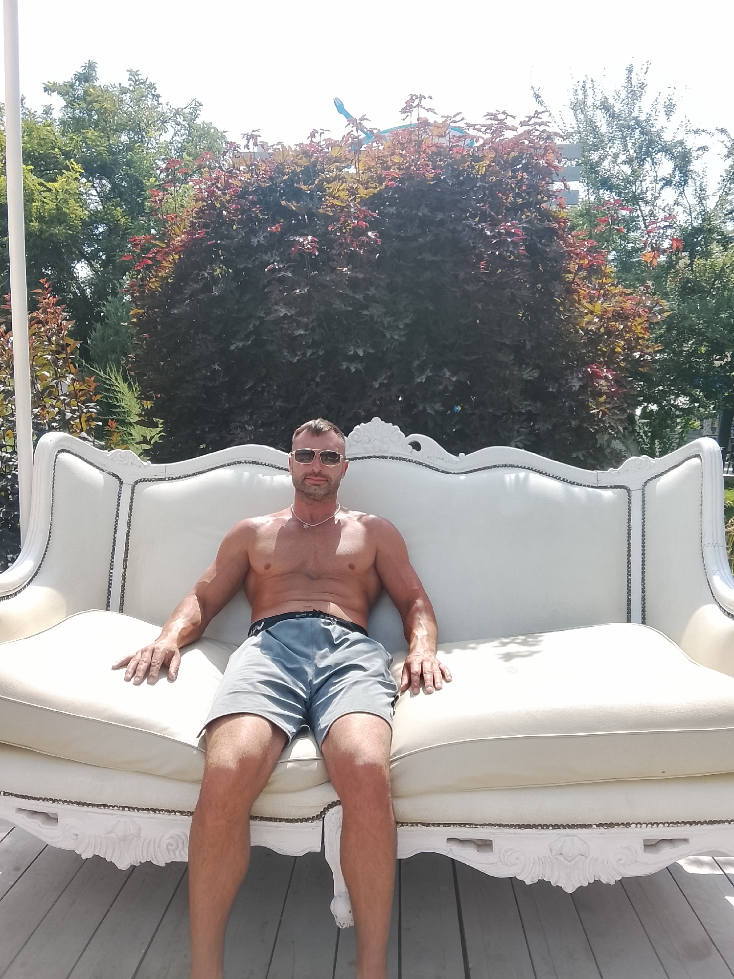 A shirtless man with short brown hair, wearing sunglasses and grey shorts, is sitting on a large, ornate white sofa. The sofa has decorative carvings and is positioned outdoors on a wooden deck. Behind the sofa, there are lush green trees and shrubs with some red foliage. The background suggests a sunny day, with bright light and shadows visible on the sofa and deck. There is no specific indication of the location being in Chișinău, Moldova.