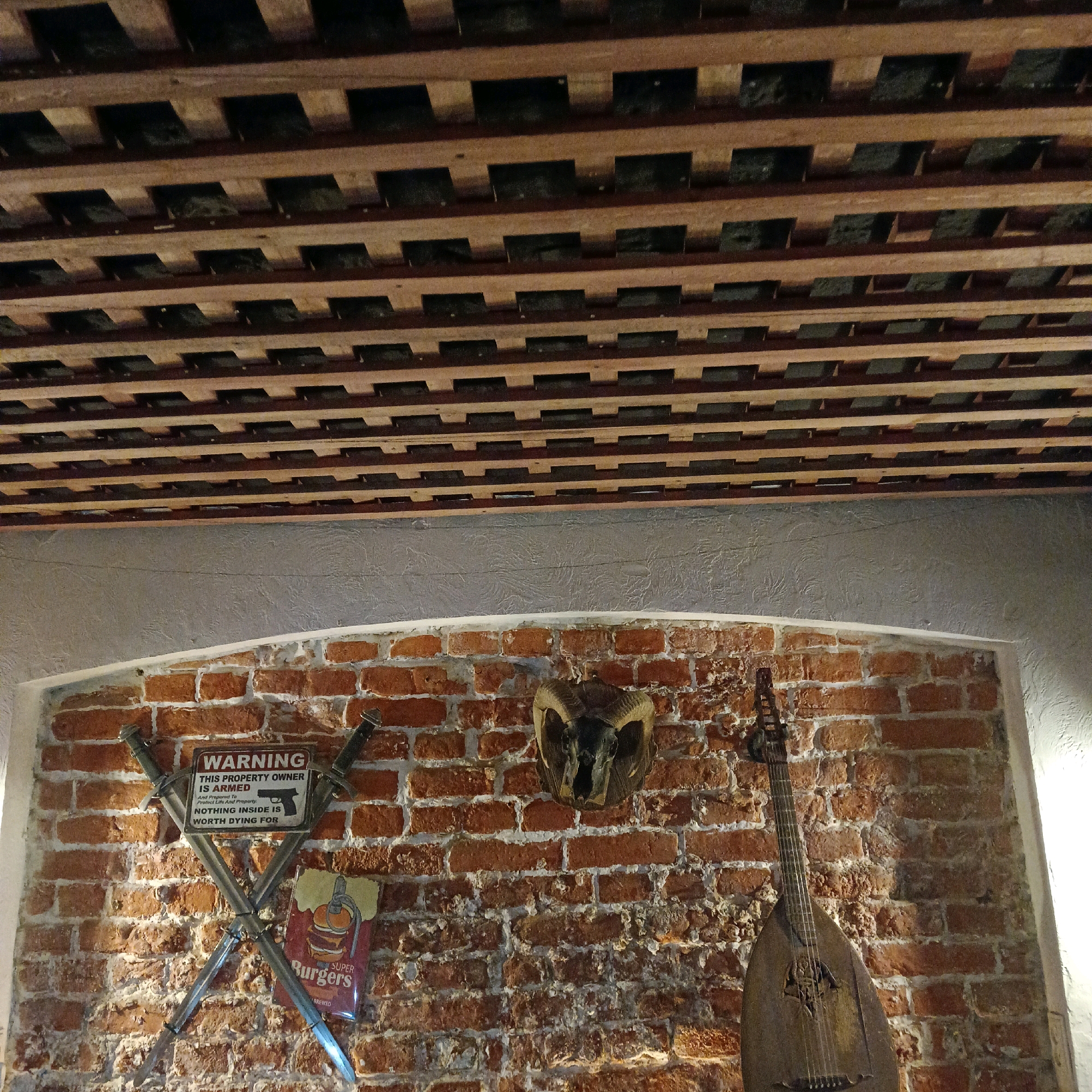 The image shows an interior wall made of red brick, decorated with various objects. At the top of the image, a wooden slatted ceiling is visible. On the brick wall, there are two crossed swords with a warning sign between them that reads "WARNING THIS PROPERTY OWNER IS ARMED And Prepared To Defend His Life And Property. NOTHING INSIDE IS WORTH DYING FOR." Below the warning sign and to the left, there is a vintage-style metal sign depicting a hamburger with the text "SUPER Burgers." Centered on the wall is a mounted ram's skull with horns. To the right of the ram's skull, a wooden stringed instrument, resembling a lute or balalaika, hangs vertically. The lighting suggests an indoor setting, possibly a bar or restaurant, with a warm and rustic ambiance. No people are present in the image. This scene is located in the city of Samara, Russia, at a establishment called "The Bard" which is a pub and restaurant.