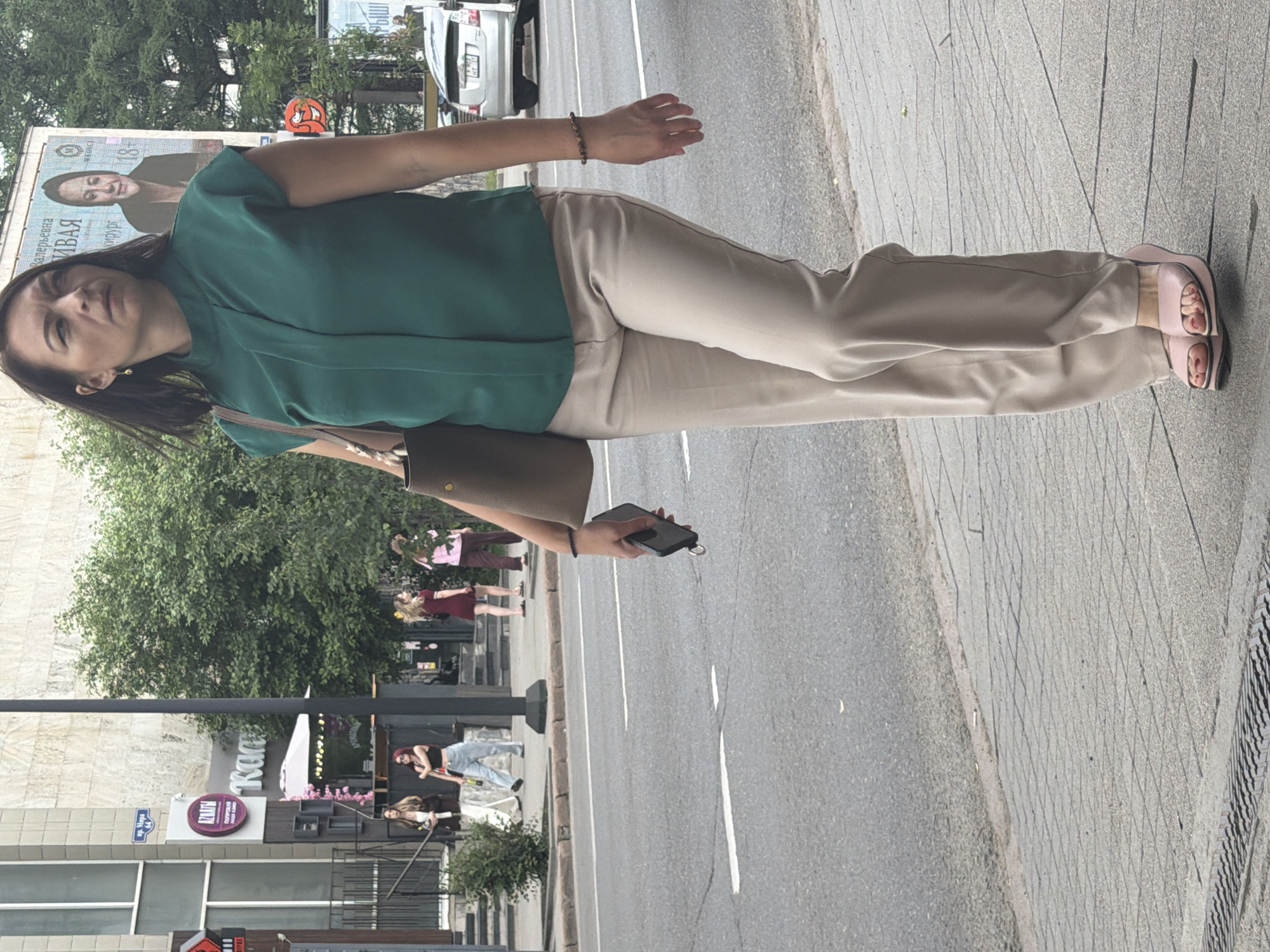 A woman with dark hair and bangs, wearing a green blouse and beige pants, walks down a paved sidewalk next to a street. She is holding a black smartphone in her right hand. Her left arm is bent, and she has a bracelet on her wrist. She wears pink slide sandals.
In the background, a large billboard features a black and white image of a woman. To the left of the billboard, a silver car is parked. Behind the woman, several other people are visible, some walking and others standing. Further in the background, trees and buildings can be seen, suggesting an urban environment. One building has a sign with Cyrillic text.
The scene appears to be outdoors during the daytime, under cloudy skies. The overall mood is casual and everyday.
Visible text: On the billboard, Cyrillic text is partially visible, including "ВАЯ" and "ири.р". A sign on a building also shows Cyrillic text, including "КЛИНИ".