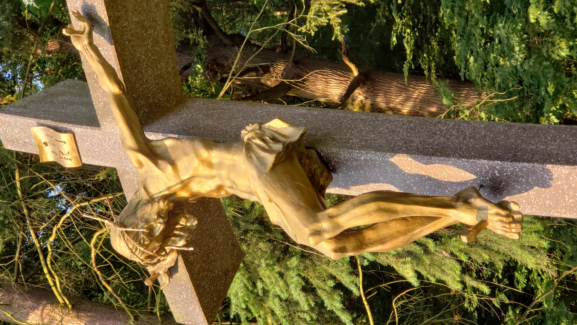 The image shows a close-up, low-angle shot of a golden statue of Jesus Christ on a cross, set against a backdrop of trees. The statue depicts Jesus with a crown of thorns, a loincloth, and his arms outstretched. His body is emaciated, and nails are visible piercing his feet. The cross itself is made of granite and has a plaque with inscribed letters "INRI" at the top. The lighting suggests it is either early morning or late afternoon, with golden sunlight illuminating the statue and casting shadows. The surrounding environment is a forest with lush green foliage and textured tree trunks.
Specifically, this statue is located at the Kalvária (Calvary) complex in Prešov, Slovakia. The complex is a significant religious and historical site, featuring a series of chapels and crosses on a hill overlooking the city. This particular cross is one of the prominent features within the Kalvária.