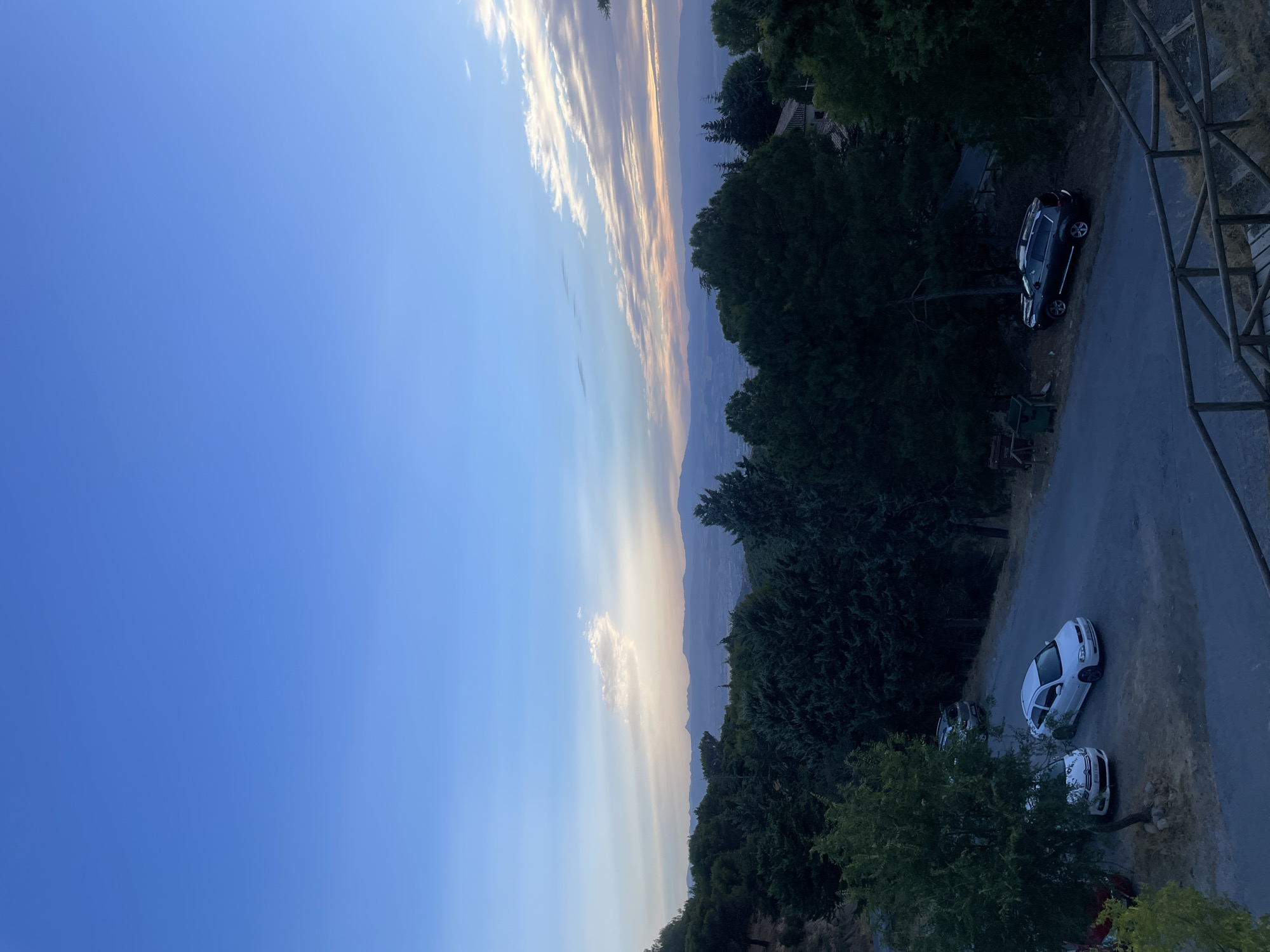 The image depicts a scenic overlook in Víznar, Spain, characterized by a panoramic view of rolling hills and distant mountains under a twilight sky. The foreground features a paved road with several parked vehicles: a white hatchback, a white sedan, and a dark SUV. Dense evergreen trees line the road and the slopes, suggesting a rural or mountainous setting. A wooden railing and steps are visible at the bottom right, indicating a designated viewing area or pathway. The sky transitions from a clear blue overhead to soft hues of orange and pink near the horizon, with wispy clouds adding texture to the scene. The overall atmosphere is serene and tranquil, capturing the quiet beauty of the Spanish countryside at dusk.