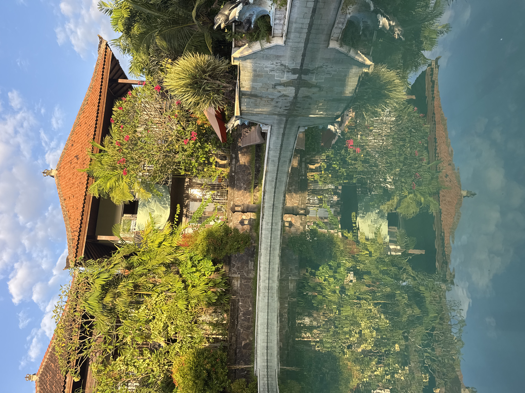 This media file captures a serene outdoor scene, likely in a tropical resort or traditional Balinese setting. The dominant subject is a two-story traditional Balinese-style building with a reddish-brown tiled roof, ornate gables, and exposed wooden beams, situated on the left side of the frame. Lush, vibrant green vegetation, including palm trees and various flowering bushes with small red blooms, surrounds and partially covers the building, indicating a rich garden environment.
A calm body of water, possibly a pool or pond, occupies the right side of the image, acting as a perfect mirror. It vividly reflects the building, the sky, and the surrounding foliage, creating an almost symmetrical composition. A stone or concrete pathway, appearing to be a bridge, extends across this water body from the foreground towards the background, connecting two land masses. Near the pathway on the left, intricate stone sculptures are visible, one resembling an animal head, possibly a mythical creature, and an arched stone structure, adding to the decorative elements.
The time of day appears to be late morning or early afternoon, characterized by bright, warm sunlight illuminating the scene and a clear blue sky with scattered white clouds visible in the upper left. The water's surface is exceptionally still, resulting in a crisp and undistorted reflection. The overall atmosphere is one of tranquility and natural beauty, heavily influenced by Balinese architecture and tropical landscaping. No specific activity or event is taking place, and there is no visible text in the image. The location cues strongly align with the given context of Kuta Selatan, Indonesia.