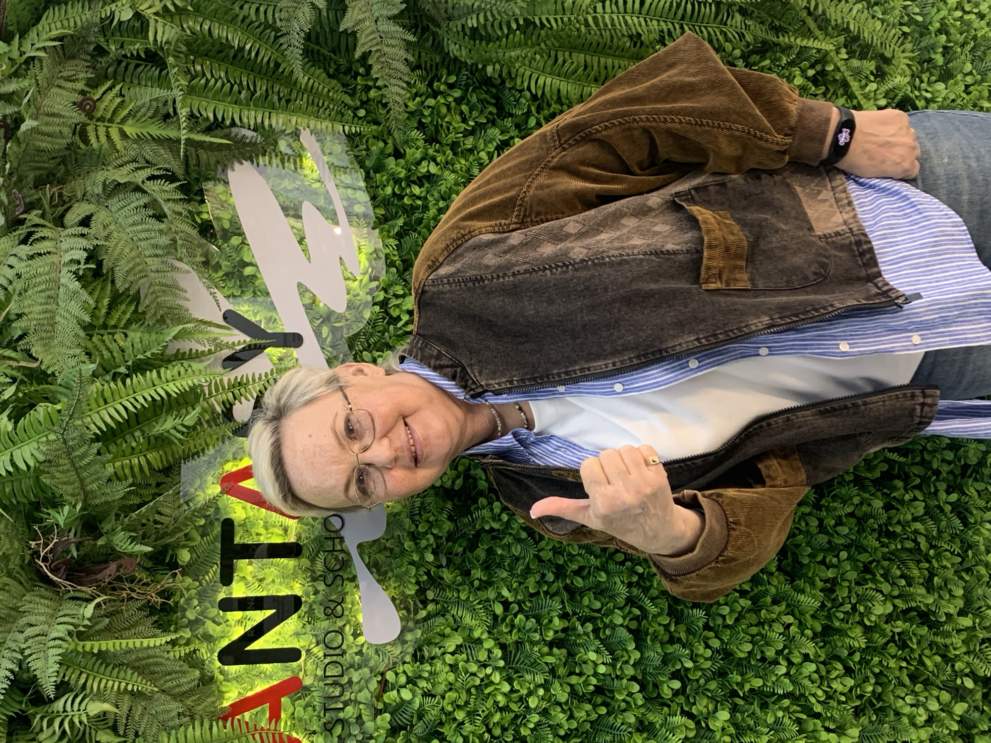 A smiling woman with short blonde hair and glasses lies on a bed of artificial greenery. She wears a blue and white striped shirt underneath a brown and black corduroy jacket, jeans, and a black fitness tracker on her left wrist. She gives a thumbs-up with her right hand, on which she wears a gold ring. In the background, "KONT STUDIO & SCHO" is displayed vertically in black and red, with a stylized white spoon image. The artificial greenery includes both fern-like plants and small, round leaves. Based on the signage and context, the location is likely in Chaikovsky, Russia.