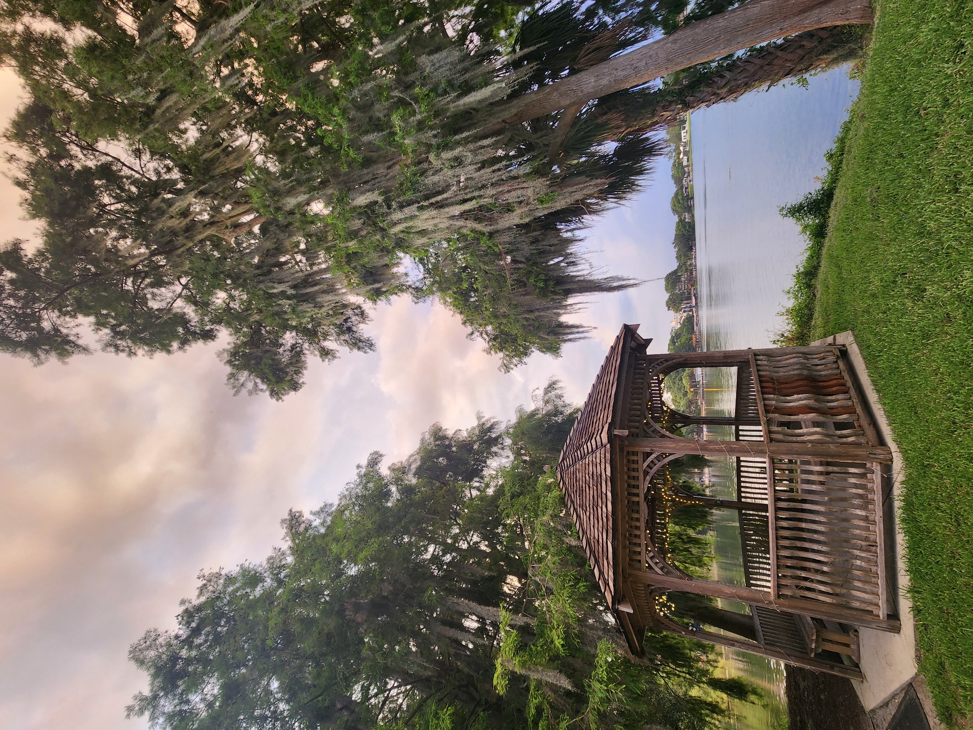 A wooden gazebo with a shingled roof, decorated with string lights, sits on a concrete base next to a grassy area. The gazebo faces a calm body of water, likely a lake, with trees draped in Spanish moss framing the scene on the left and right. The water reflects the overcast sky, which is a mix of pale pink, orange, and gray. In the distance, across the lake, is a treeline with scattered houses. The image appears to be taken at dusk or during a cloudy sunset, as indicated by the soft lighting and muted colors. The location is likely along the waterfront in Winter Park, Florida, given the characteristic vegetation and architecture.