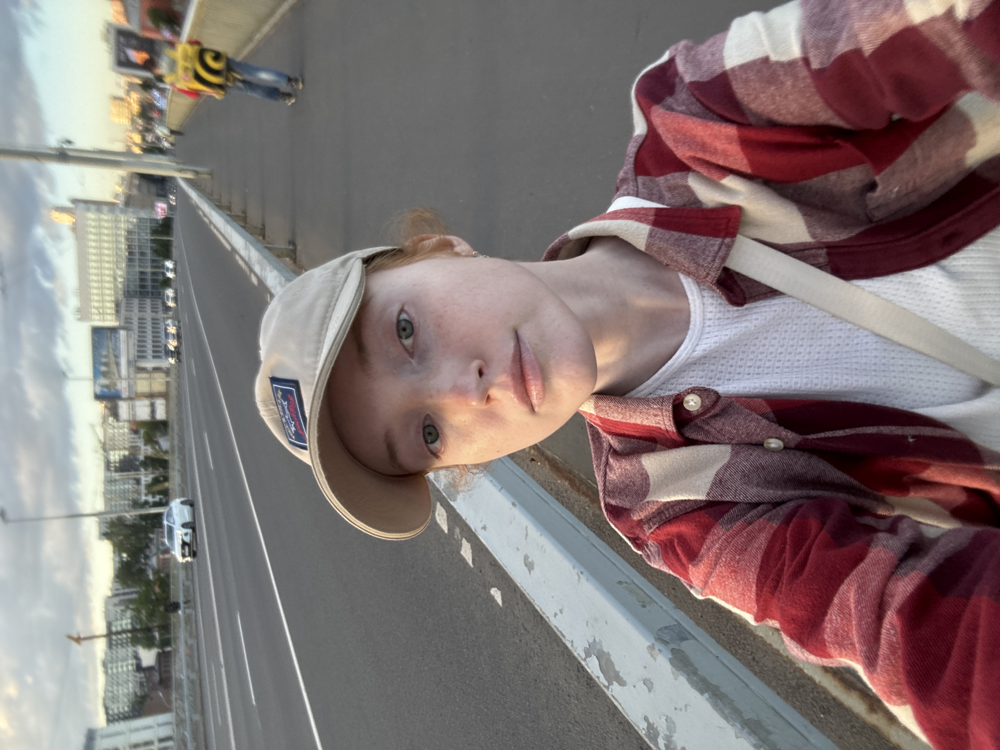 The image is a selfie taken by a young person with red hair, wearing a tan baseball cap and a red and white plaid shirt over a white waffle-knit shirt. The person has freckles and is looking towards the camera with a neutral expression. The background suggests an urban environment, likely a bridge, with cars driving on a road. In the distance, buildings and streetlights are visible. The sky appears overcast, indicating it might be late afternoon or early evening. A person in a yellow and black vest is visible in the far background, walking on the sidewalk of the bridge. There is a visible white strap across the person's chest, possibly from a bag. A small logo is visible on the side of the baseball cap. The image is rotated 90 degrees clockwise.