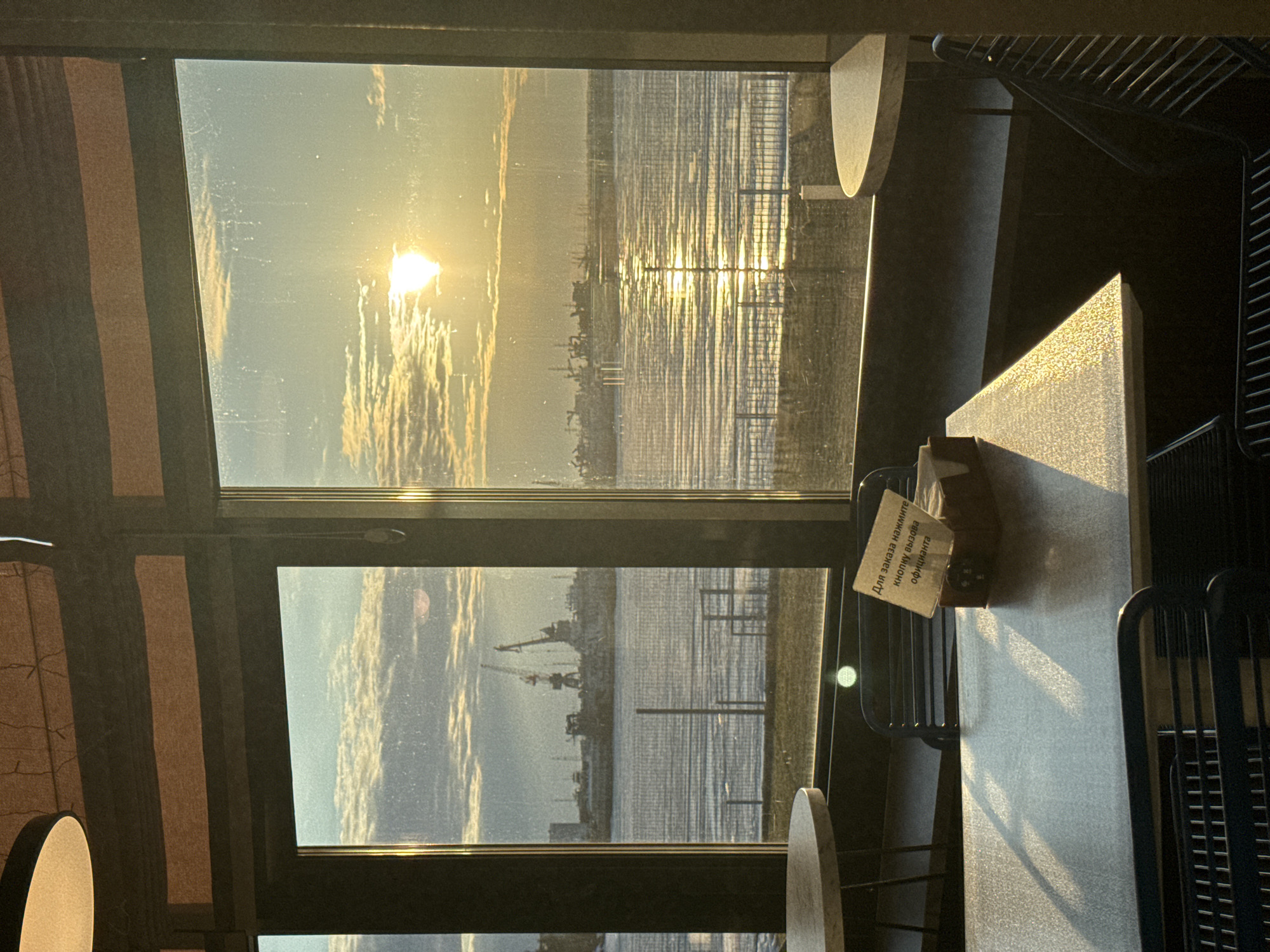The image shows an interior view through a window overlooking a body of water with industrial structures on the opposite shore. A sign is visible on the table. Based on the sign's language (Russian) and the industrial structures on the water, the location may be Chaikovsky, Russia. The sky is bright, possibly indicating sunrise or sunset. The scene is captured from inside a building, possibly a restaurant or cafe, as evidenced by the table and chairs. No people are visible in the image.