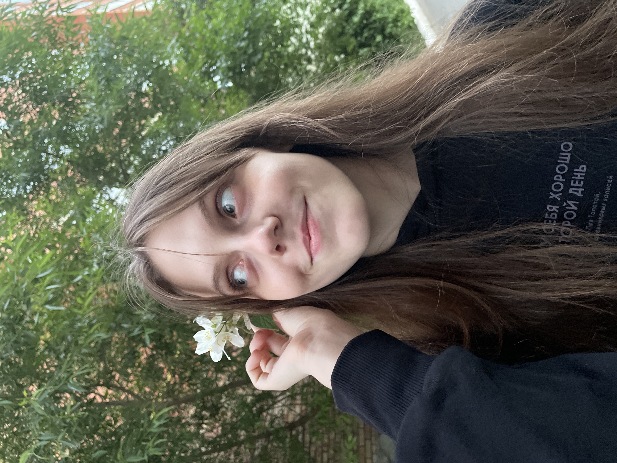 In a medium shot, a young woman with long brown hair is pictured from the chest up, holding a white flower near her head. She is looking to her right with a slight smile. She is wearing a black hooded sweatshirt with white text on it. The background consists of green trees and a building, suggesting an outdoor setting. The image is taken in Ufa, Russia.
