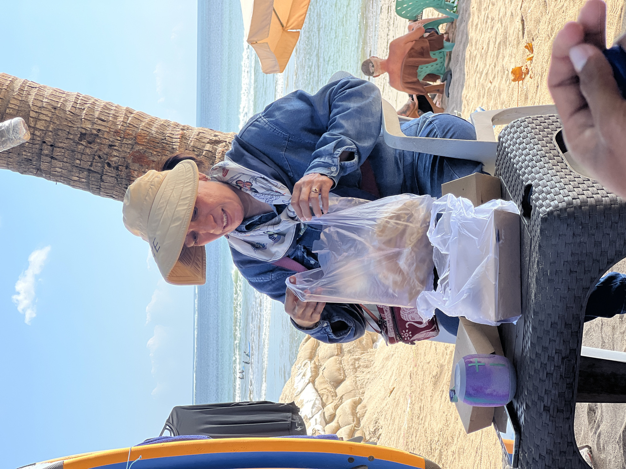 A woman wearing a wide-brimmed hat and denim clothing sits on a plastic chair on a sandy beach in Kuta, Indonesia. She is looking down at a clear plastic bag filled with food, which she is holding with both hands. In front of her, on a woven plastic table, are two cardboard food containers, a mobile phone, and a portable speaker. To her left, a surfboard leans against a pile of sandbags. In the background, the ocean stretches out under a blue sky with scattered clouds. Two people are visible in the water, one on a surfboard. Further down the beach, a person sits on a chair under a yellow umbrella, and another person is seated in a green chair. The scene appears to be a casual outing at the beach on a sunny day.