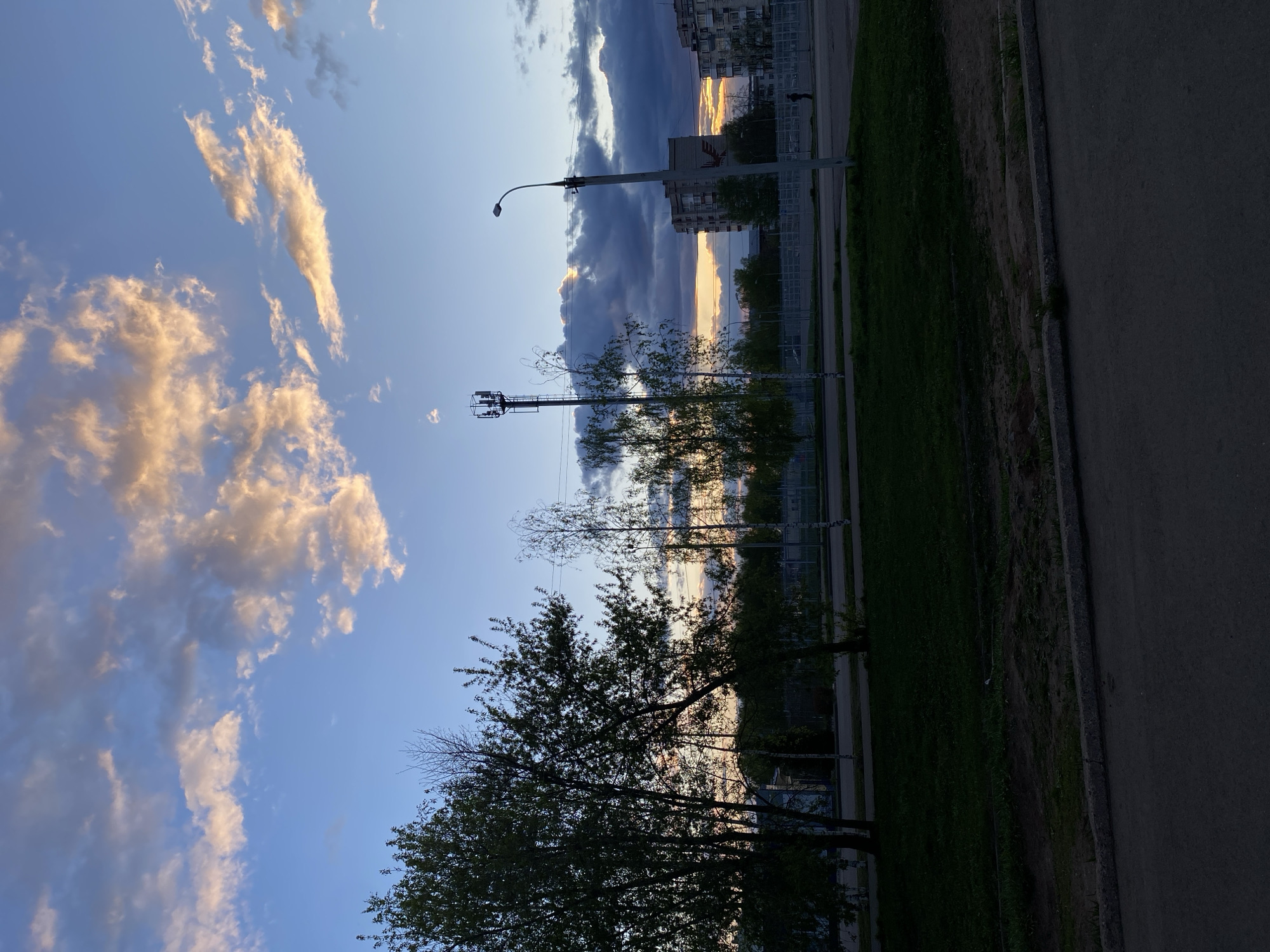 The image depicts an urban scene likely in Votkinsk, Russia, judging by architectural styles possibly unique to the region. A street runs along the right side of the frame, with a grassy area and a chain-link fence bordering it. Trees are scattered along the fence line. In the background, buildings are visible, silhouetted against the sky at sunset or sunrise. Light poles and communications towers stand near the buildings. The sky is partly cloudy. The image suggests early evening or morning hours based on the sky's brightness. A single person is walking along the street.