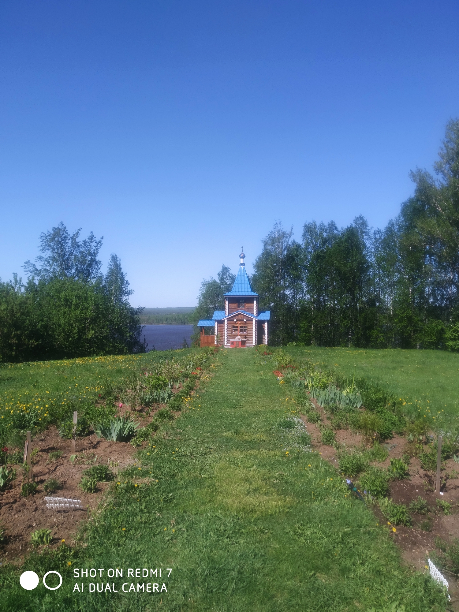 The image shows a wooden church with a blue roof in a rural setting. A grassy path leads to the church. There are flowerbeds on either side of the path. In the background is a body of water and trees. The sky is clear and blue. The location is Ochansk, Russia.
