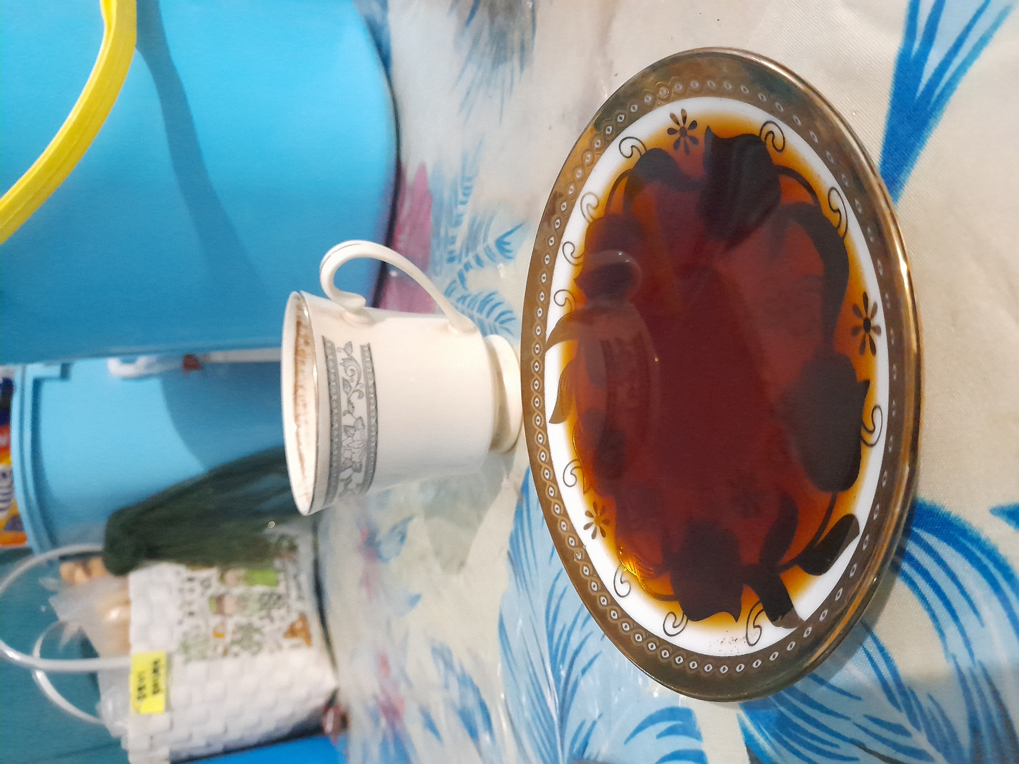 A tea cup and saucer rest on a blue and white floral tablecloth. The saucer contains a dark liquid. Behind the cup and saucer, a blue plastic container is visible. The image was taken in Gelam, Indonesia.