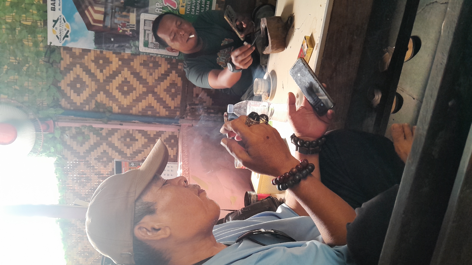 In Balun, Indonesia, two men are seated at a table outdoors. The man on the left, wearing a light blue t-shirt and a beige baseball cap, is in the foreground. He has dark hair, is looking down, and is holding a cigarette in his right hand, which is also adorned with several bracelets on his wrist. A cloud of smoke emanates from the cigarette. His left hand is holding a black smartphone.
The man on the right, wearing a dark green t-shirt, is in the background. He is also smoking a cigarette and looking down. He is holding a smartphone in his left hand. Between the two men are various objects on the table, including a bottle of water, a small cup, a wooden carving, and a pack of cigarettes.
The background features a woven bamboo wall with a diamond pattern and a poster for "GRAND TIARA" real estate. A ceiling fan is visible at the top of the frame. The overall lighting suggests it is daytime, with sunlight streaming in from the left.