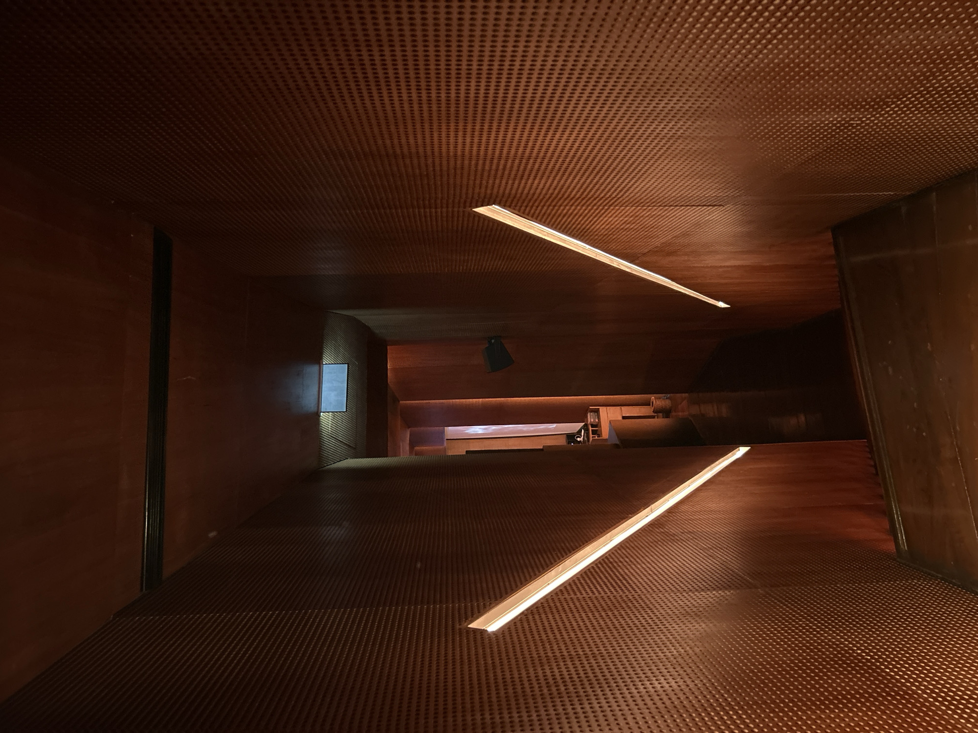 The image depicts a dimly lit interior hallway with walls and ceiling clad in dark wood panels featuring a subtle, repetitive perforation pattern. Two diagonal light fixtures, emitting a warm, white glow, are embedded in the walls on either side of the hallway, creating sharp, geometric accents. The hallway appears to descend, with visible wooden steps at the bottom of the frame. In the distance, the hallway opens into a slightly brighter area with more wooden columns and a glimpse of what might be a reception desk or counter. The overall ambiance is modern and sophisticated, with a focus on warm tones and geometric design. This scene is located in the city of North Kuta, Indonesia.