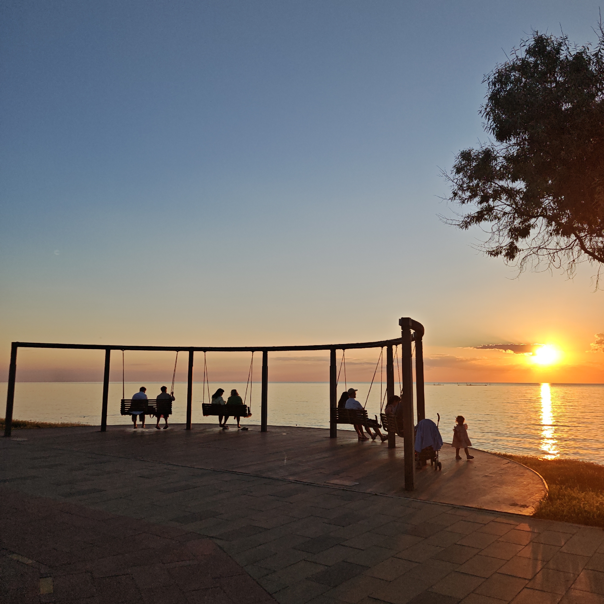 An outdoor, eye-level, full shot captures a silhouetted scene during sunset at the edge of a body of water. In the foreground, a paved walkway with a tiled pattern stretches horizontally across the frame. A series of swings are set up on a wooden platform adjacent to the walkway, facing the water.
On the swings, multiple individuals are silhouetted against the bright sky and water. On the leftmost swing, two figures are seated. The next swing to the right has three figures seated, one appearing to be a child. The third swing from the left has two figures seated, one with a hat. The fourth swing from the left has two figures seated.
To the right of the swings, a stroller is visible, and a young child, also in silhouette, is walking away from the viewer towards the water. A tree with dark foliage extends into the top right corner of the frame, its branches silhouetted against the sky.
The sky transitions from a light blue at the top to a gradient of soft oranges and yellows near the horizon, indicating sunset. The sun itself is a bright, intense orb, partially obscured by a small cloud, casting a radiant glow and a shimmering reflection on the rippling water. The water extends to the horizon, meeting the sky in a soft blend of colors. The overall mood is peaceful and serene.