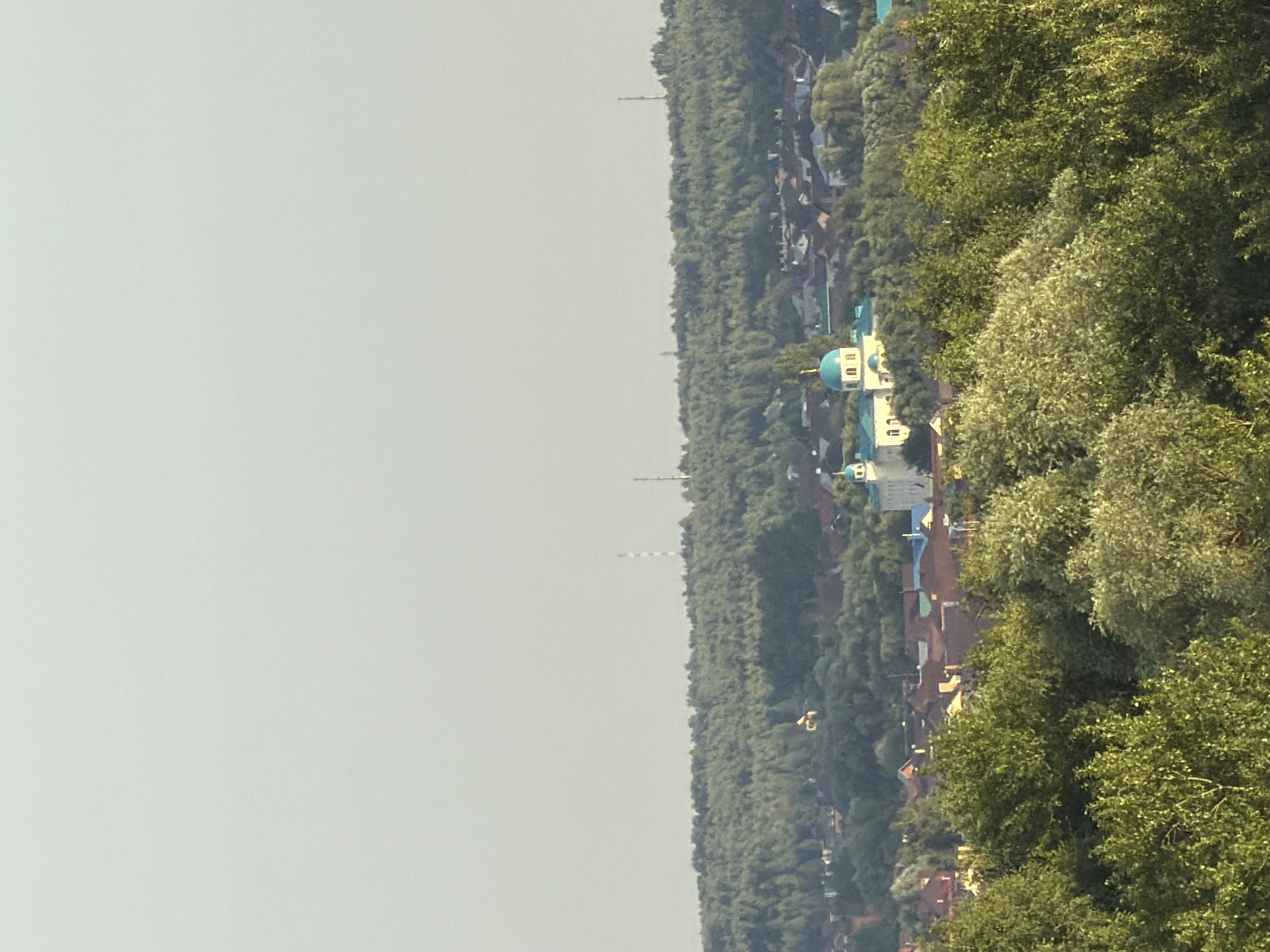 The image shows a landscape view of a city from a distance, with a clear sky and a distant treeline. In the mid-ground, there is a cluster of buildings, prominently featuring a white church with turquoise domes. The church appears to be the Church of the Assumption of the Blessed Virgin Mary, located in Kazan, Russia. Several other buildings with red roofs are visible in the vicinity. The foreground is dominated by lush green trees, suggesting a natural setting overlooking the urban area. The overall scene depicts a peaceful, somewhat hazy, daytime view of a town or city.