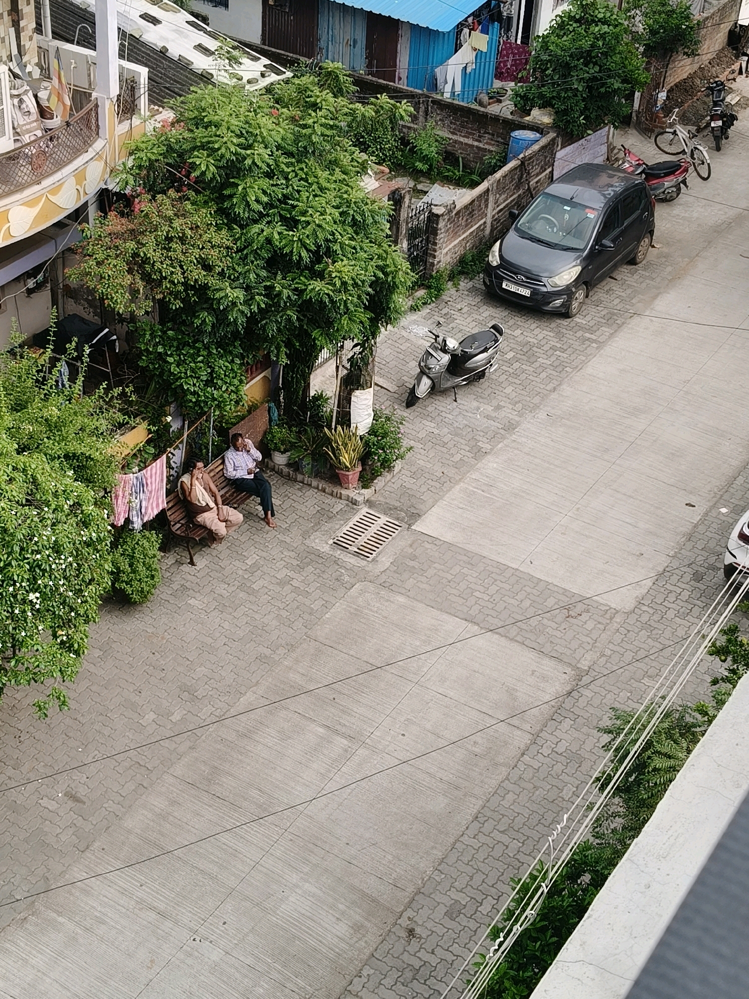 The image is an aerial view of a street in Nagpur, India. In the foreground, two men are sitting on a park bench. The man on the left is wearing a light-colored shirt and pants, while the man on the right is wearing a blue and white checkered shirt and dark pants. They appear to be talking on their phones. Behind them, a tree with green leaves and white flowers is in bloom. A clothesline with striped fabric is hanging from a pole next to the tree.
To the right of the men and the tree, there is a dark gray car parked on the street, which is paved with interlocking concrete blocks. Further down the street, a white car is visible. Several motorbikes are also parked along the street. The background shows buildings with pitched roofs, some of which are made of corrugated metal. A blue tarp is stretched over a section of one of the buildings. The overall impression is of a residential area in a city with a mix of old and new architecture.