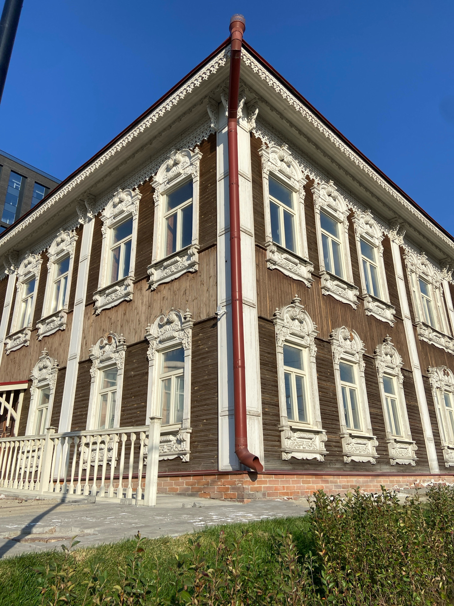 The image shows a wooden building with ornate white trim around the windows and along the roofline. The building has a red downspout running down the side and a white railing in front. The building is located in Novosibirsk, Russia, in a clear blue sky on a sunny day. In the background, a modern building with dark windows can be seen. The foreground shows a patch of green grass. The building appears to be historic architecture.
