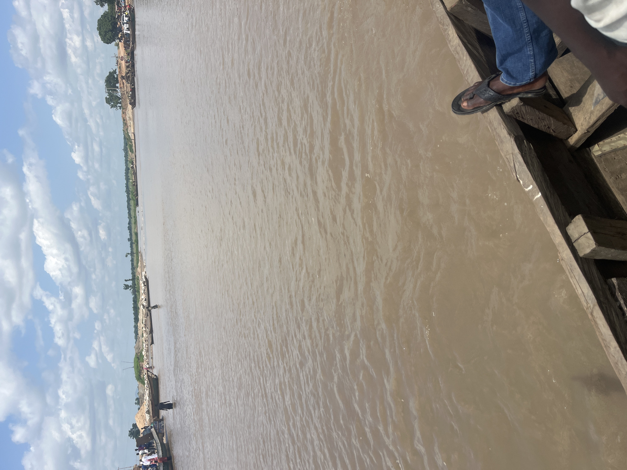 A wide, muddy river flows through a scene of human activity in Nemnei, Nigeria. Several wooden boats are visible, some carrying passengers and others appearing to be used for transport of goods, possibly sand or gravel, given the piles on the riverbank. People are seen wading in the shallow water near the bank, and some are standing on the boats. The riverbank is lined with sand deposits and sparse vegetation, with trees and buildings visible in the distance. The sky is partly cloudy with white clouds against a blue backdrop. In the foreground, the wooden interior of a boat is visible, with a person's legs and feet, wearing blue jeans and black sandals, resting on the edge. The water appears calm with small ripples.