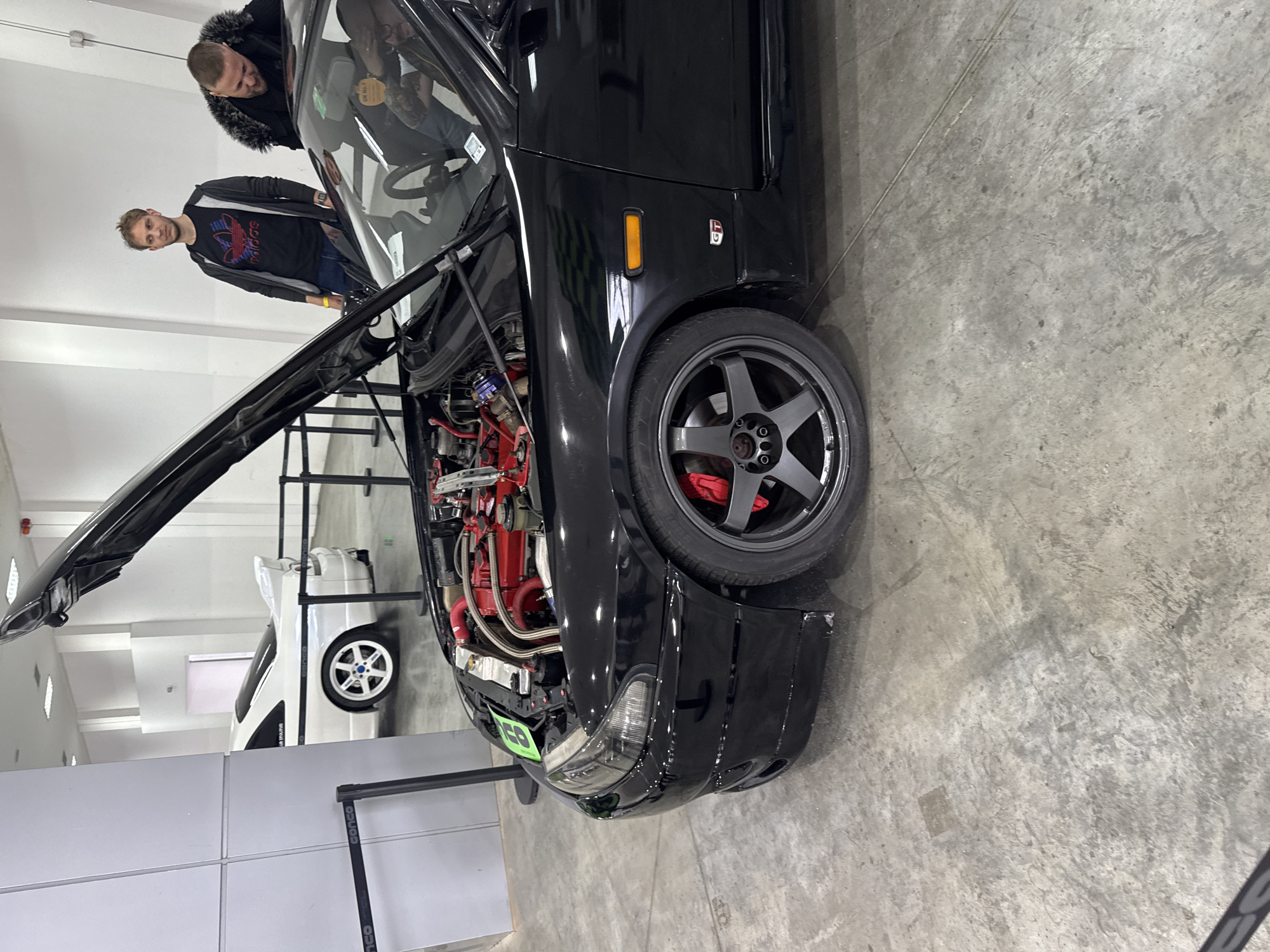 The image depicts a black Nissan Skyline R33 GT-R with its hood open, showcasing a modified engine bay with red hoses and components. The car is located indoors, possibly at an auto show, as suggested by the concrete floor and bright lighting. Two men are looking at the vehicle, one wearing a black jacket with fur trim and the other in an Adidas shirt under a black jacket. In the background, partially visible, there is a white car and partitioning barriers. The scene is likely not in Plovdiv, Bulgaria, as there is no indication of the location.