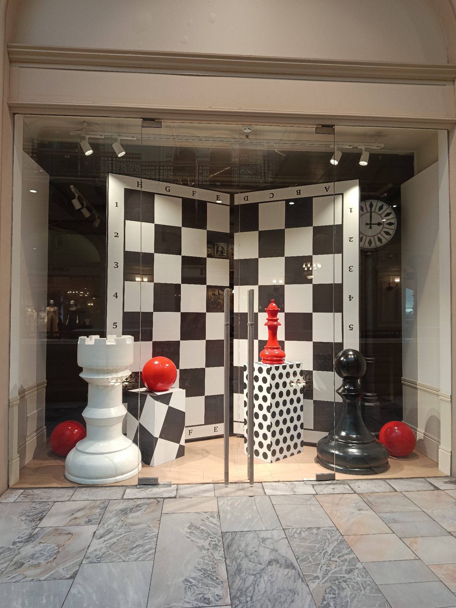 The image displays a vibrant retail window showcasing an oversized, art-themed chess setup. The scene is set indoors, likely within a shopping mall or department store, as suggested by the reflective glass, visible interior lighting fixtures, and the marble-patterned tiled floor in the foreground.
The central feature is a large, two-panel, black and white checkered backdrop resembling a chess board, angled to form a corner. This board is marked with algebraic chess notation: letters "H" through "A" are visible along the top edges, and numbers "1" through "5" run vertically down the sides, seemingly indicating a limited representation or decorative scale rather than a full board.
Prominently featured are three dramatically oversized chess pieces: a white rook positioned on the left, a striking red queen elevated on a black and white polka-dotted pedestal in the center, and a black pawn on the right. Three bright red spherical objects are strategically placed around the pieces and on a smaller checkered pedestal, adding bold accents.
Reflections on the glass reveal elements of the surrounding interior space, including a large, classic wall clock with Roman numerals on the right and faint silhouettes of other displays or mannequins on the left. Overhead track lighting illuminates the scene, highlighting the stark contrasts and playful elements of the display. The arrangement creates a sense of grandeur and whimsy, serving as an eye-catching artistic installation.