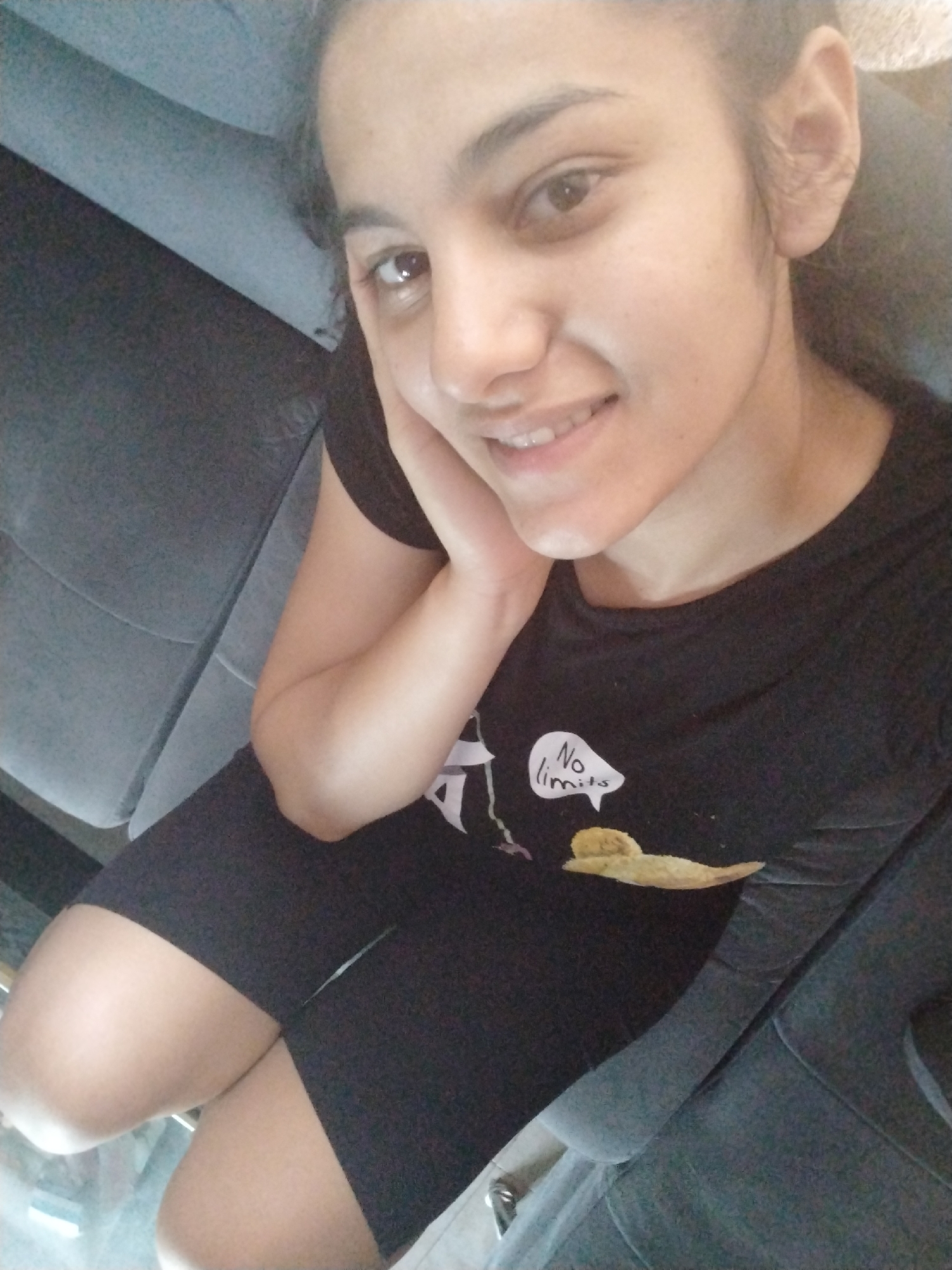 This is a selfie of a young woman sitting on a dark gray sofa. She is wearing a black t-shirt with a graphic design on it and black biker shorts. The t-shirt has a speech bubble with the words "No limits" written inside, and a drawing of a piece of food below it. The woman has dark hair and is smiling at the camera, with her hand resting on her cheek. The lighting suggests it could be daytime, and the overall mood appears relaxed and casual. The background is dominated by the sofa, with a hint of what looks like a patterned material in the upper left corner.