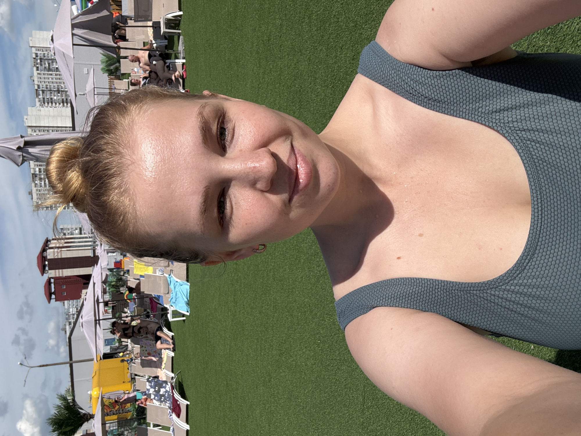 A young woman with blonde hair in a bun, wearing a dark teal swimsuit, takes a selfie on a sunny day outdoors. She is looking at the camera with a slight smile. In the background, there is a well-maintained green lawn, several lounge chairs, and some people relaxing. In the distance, there are tall buildings and a yellow food truck. The image appears to have been taken at a public outdoor venue, possibly a water park or a recreational area, within the city of Yaroslavl, Russia.