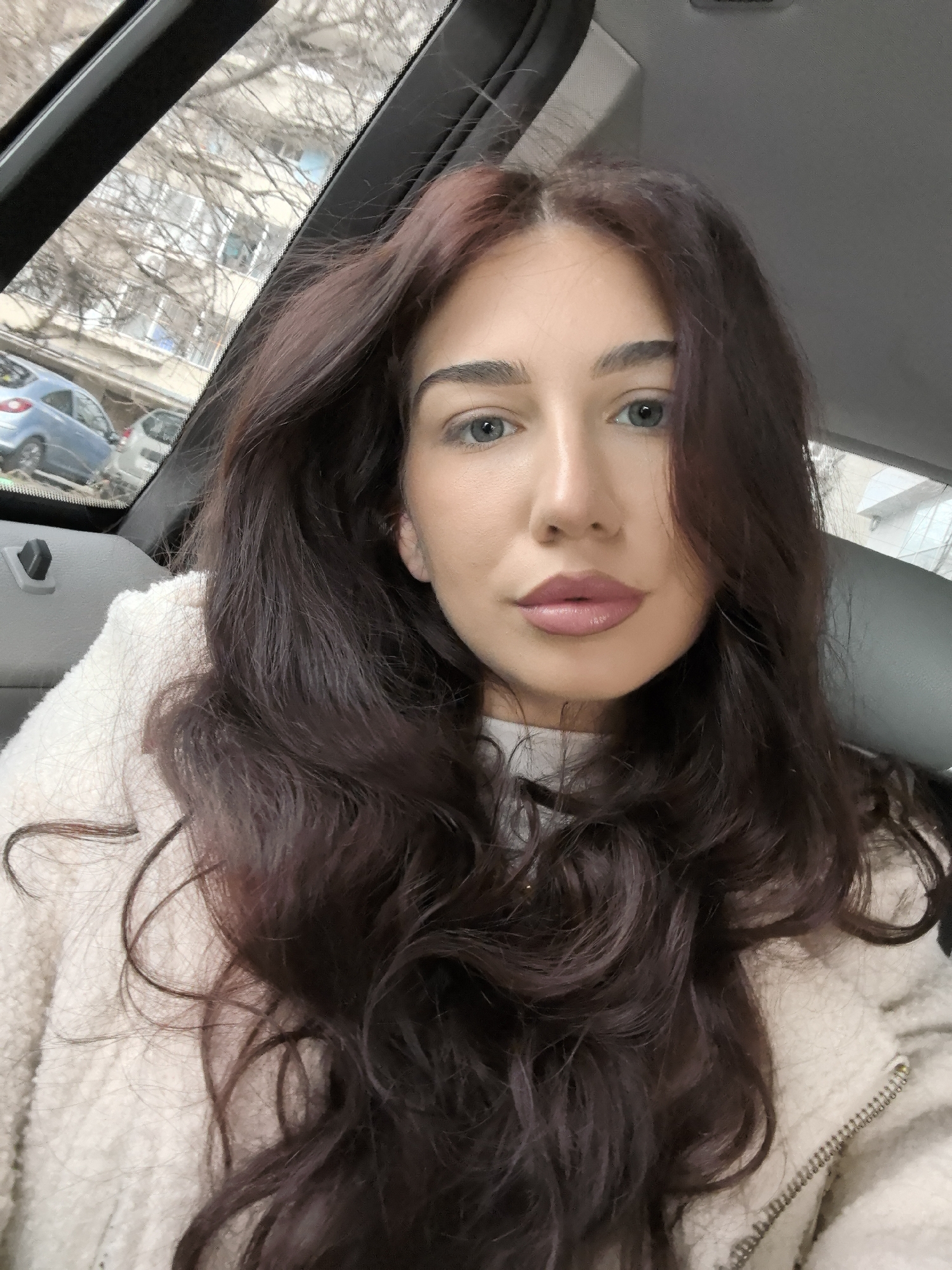 A young adult female is depicted in a close-up, frontal portrait, seated inside a vehicle. She has long, dark brown, wavy hair with subtle reddish undertones, blue-grey eyes, and wears pink lipstick. Her attire includes an off-white, textured fleece jacket with a visible zipper, over a white inner garment. The vehicle's interior shows a grey headliner and black trim around the window. Through the vehicle's window, an urban street environment is visible, consistent with the city of Stara Zagora, Bulgaria. The background elements include several parked vehicles, notably a light blue compact car, bare deciduous tree branches, and a multi-story building with light-colored facades and numerous windows.