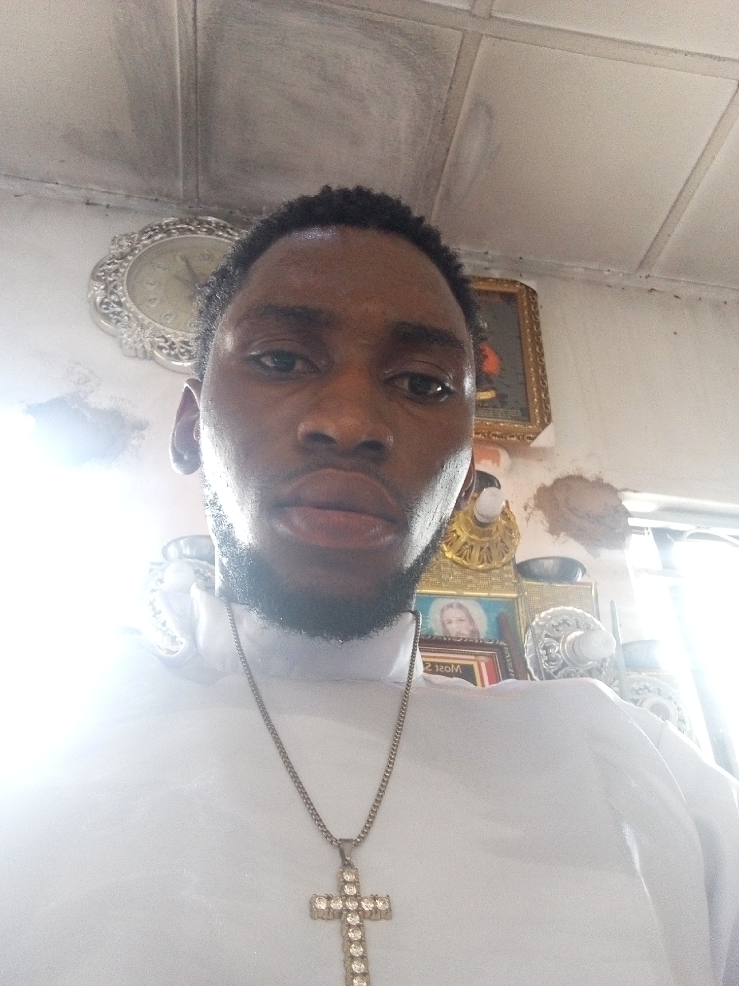 A mid-level angle shot of a dark-skinned man with short, dark hair and a beard. He is wearing a white garment and a gold chain with a cross pendant. Behind him, a wall displays various framed pictures, a clock, and decorative items. The location is unknown, city and region unspecified.