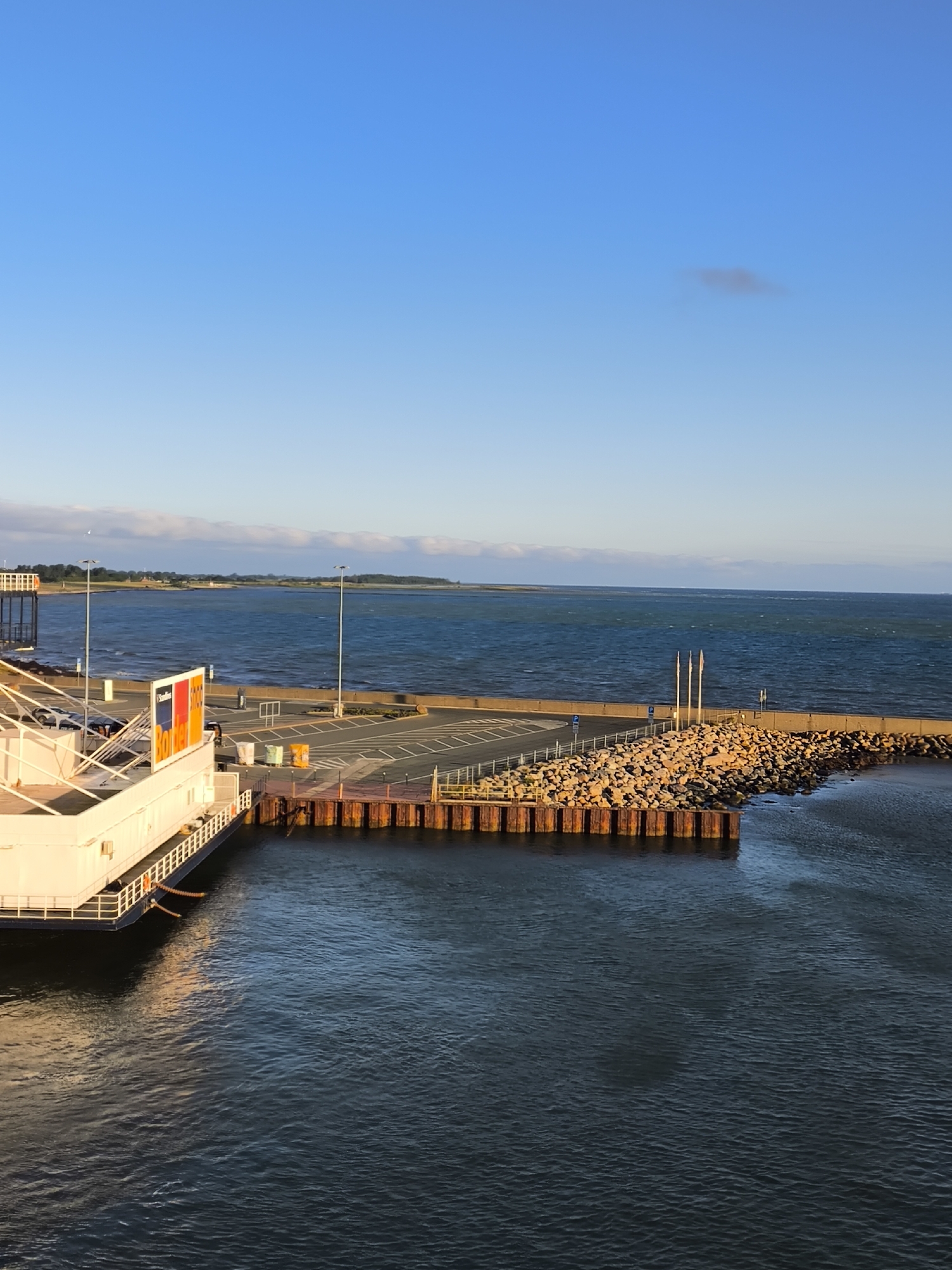 This is a vertical, eye-level shot taken from a ferry looking towards a ferry terminal on a clear day. The sky is a bright, gradient blue with a single, wispy cloud. The water is dark blue with gentle ripples. In the foreground on the left, a white ferry is docked, with its railing and some orange ropes visible. The ferry terminal has a paved area with parking spaces and some colorful bins. A breakwater made of large rocks extends into the water, with several flagpoles on top. The background shows a distant coastline with trees and buildings. The overall scene depicts a functional ferry port with a backdrop of natural scenery under a serene sky. This is likely the Puttgarden ferry terminal on the German island of Fehmarn.