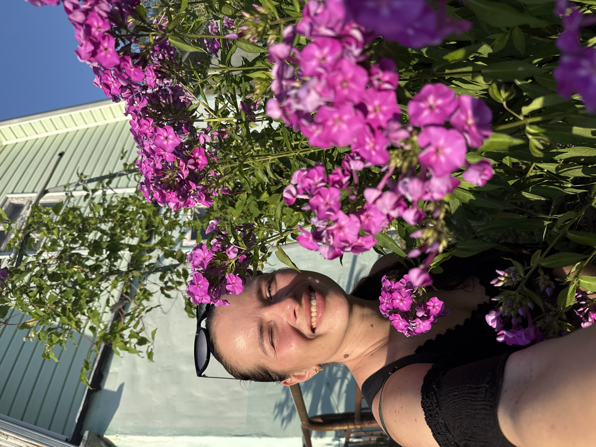 A young woman with dark hair is captured in a close-up, outdoor portrait, her face illuminated by the sun. She is smiling broadly, with her eyes closed, conveying a sense of joy and contentment. The foreground is dominated by a cluster of vibrant pink phlox flowers, framing her face and adding a splash of color to the image. She is wearing a black top and sunglasses perched on her head.
Behind the woman and the flowers, a light blue building with white trim is visible. The building's siding appears to be corrugated metal. The sky is a clear, bright blue, suggesting fair weather and likely daytime. The overall scene suggests a relaxed and pleasant outdoor setting, possibly a garden or yard. The orientation of the image is rotated 90 degrees counter-clockwise.