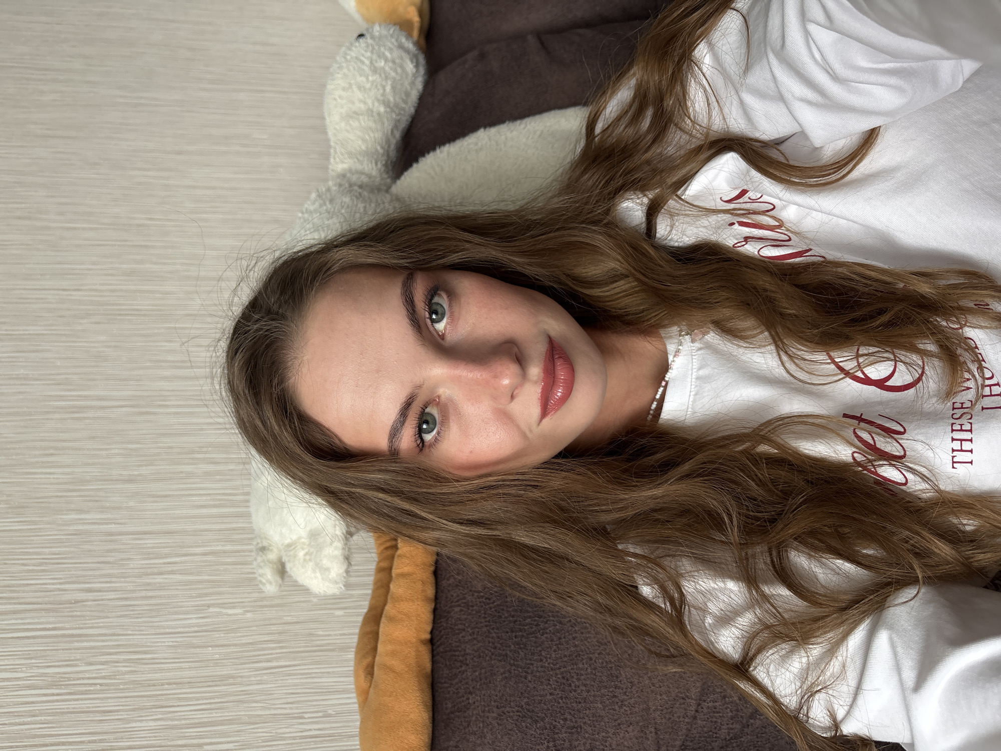 An indoor, chest-up selfie depicts a young adult female looking directly at the camera with a slight smile. She has long, wavy, light brown hair, parted in the middle and flowing over her shoulders and chest. Her eyes are hazel/green, and her lips are full with a glossy, berry-toned application. Her fair complexion shows a natural flush on her cheeks and nose. She is wearing a white long-sleeved crew-neck top adorned with red script lettering, partially visible as 'set &' and 'THESE W'. A white beaded necklace with a small pink charm is visible around her neck. She is seated on a dark brown, textured couch. Behind her, resting on the couch back, are two plush items: a segment of a long, orange-brown stuffed object on the left, and a white, fluffy, sheep-like stuffed animal with black facial features on the right. The background wall features light beige wallpaper with a subtle vertical stripe pattern. The scene is located in the city of Perm, Russia.
