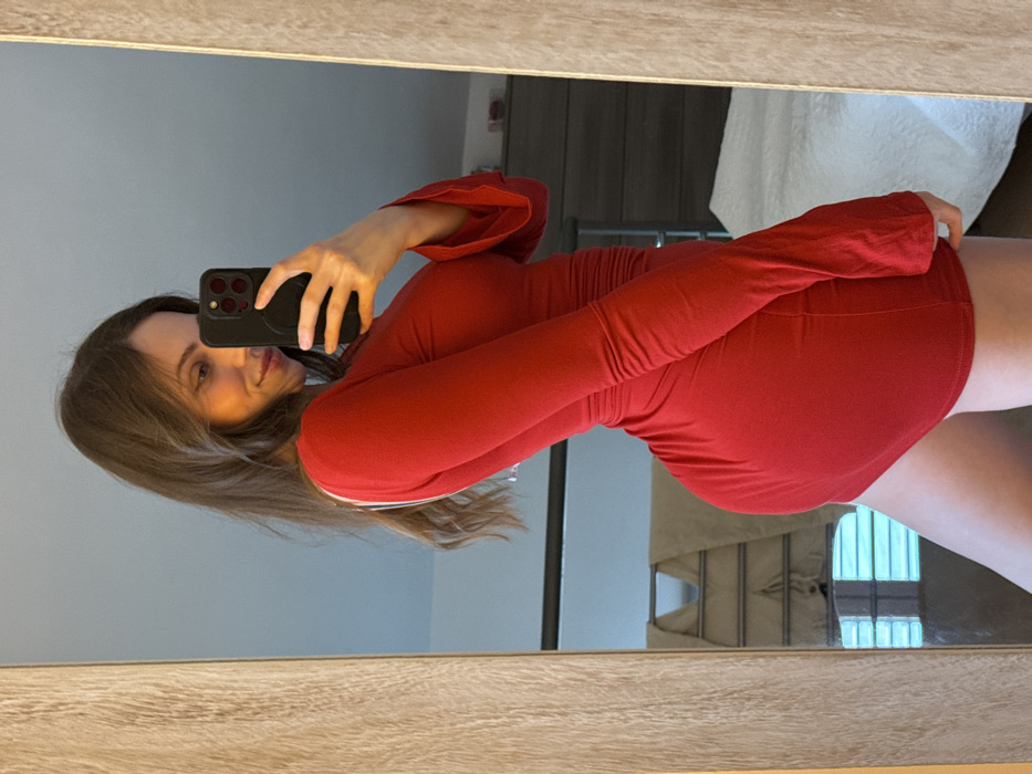 A female individual is depicted in a full-length mirror selfie taken within a private residence in Cava de' Tirreni, Italy. The subject, a woman with long, dark brown hair, is wearing a form-fitting, long-sleeved, v-neck red mini-dress. A white strap is visible over her left shoulder. She is angled slightly to her left, looking over her right shoulder towards the camera with a subtle smile. Her right hand holds a dark-colored smartphone, equipped with a circular grip accessory on its back, utilizing the mirror to capture her reflection.
The mirror, framed in light-toned wood, reflects elements of the room interior. Visible in the background reflection are a bed with white bedding on the right, a dark-colored dresser or chest of drawers in the upper-right quadrant, and a silver-colored, collapsible clothes drying rack in the middle-left, with a light-colored towel draped over it. The visible wall surfaces are a light blue or grey color, indicating an indoor, likely bedroom, environment.
