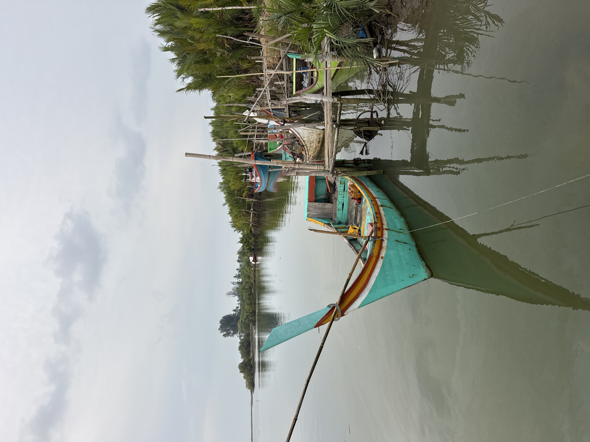 The image depicts a calm river scene in Geuleudah, Indonesia, with several traditional wooden fishing boats moored along a wooden pier. The foreground features a prominent turquoise and yellow boat with a distinct pointed bow, its reflection clearly visible in the still water. Behind it, other boats, including a blue and white one and a green one, are docked. The pier is constructed of rough-hewn wooden posts and planks, with some fishing gear and debris scattered around. Lush green palm trees line the riverbank on the right, creating a dense, tropical backdrop. The sky is overcast with soft, gray clouds, and the overall lighting is diffused, suggesting a tranquil, possibly humid, environment typical of a coastal or riverine area in Indonesia. There are no people visible in the image.
