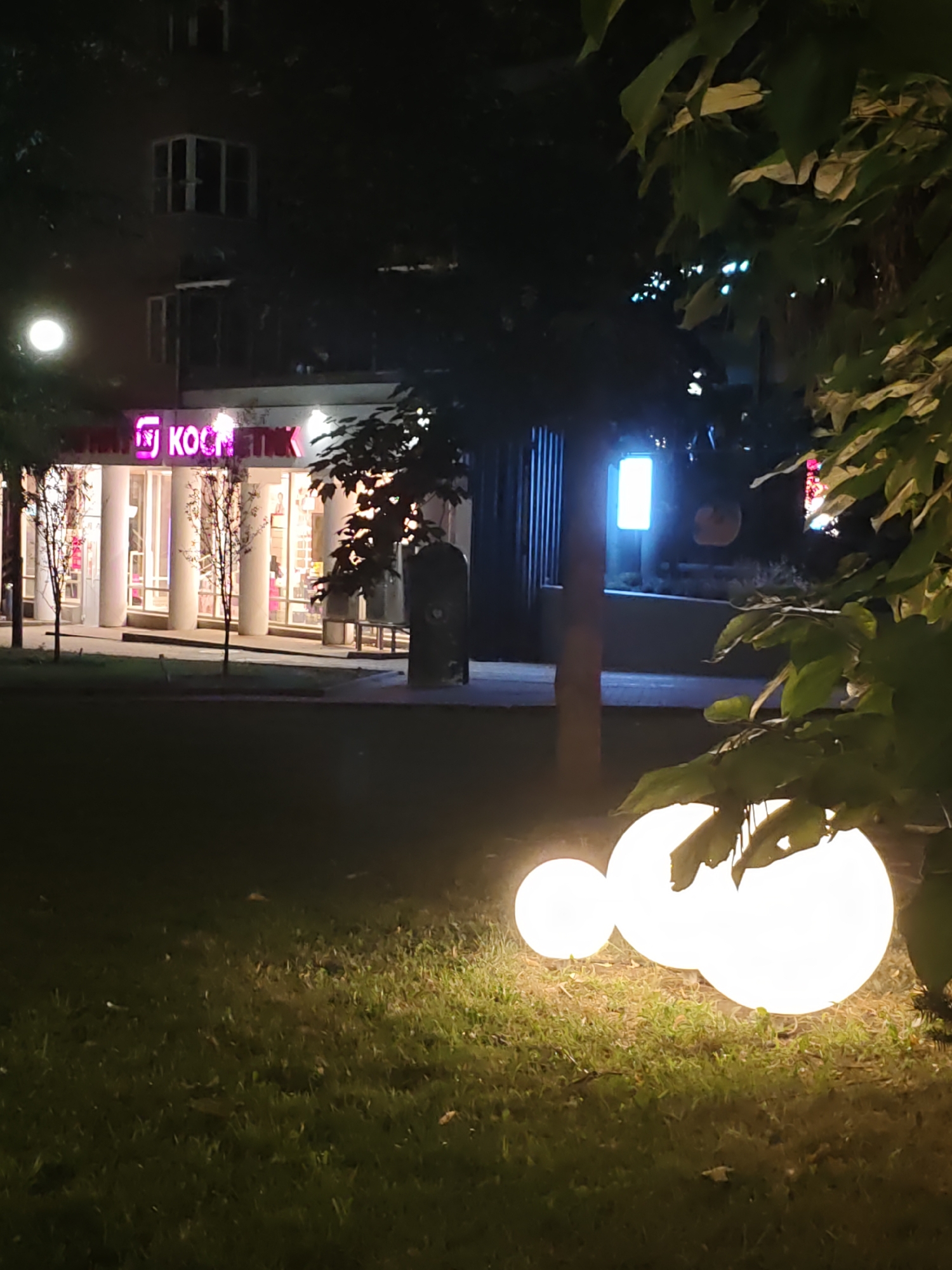 This image depicts a nighttime scene in Rostov-on-Don, Russia. In the foreground, three luminous spherical lights are placed on a grassy area. Behind them, a building with large glass windows is visible, featuring a prominent sign with pink lettering that reads "КОСМЕТИК" (Cosmetic). The building appears to be a commercial establishment, possibly a store or salon. Trees with dark foliage are present, some casting shadows and framing the scene. Other faint lights and architectural elements suggest an urban environment.