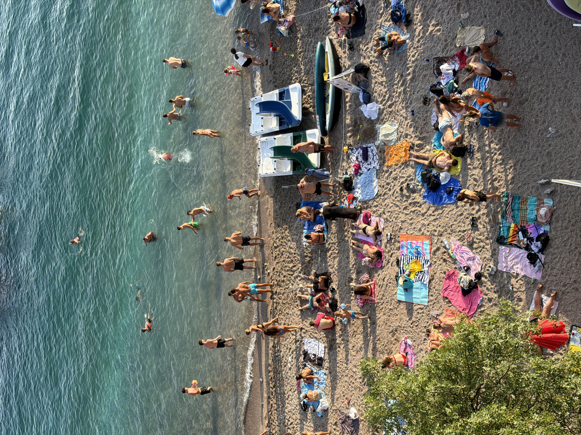 The aerial view captures a bustling beach scene in Boreti, Montenegro, with numerous individuals enjoying the clear turquoise waters of the Adriatic Sea and the sandy shore. Several people are swimming and wading in the sea, some playing with a ball. On the beach, many sunbathers are scattered across towels and blankets, some reading or socializing, while others are relaxing or sleeping. A group of children are playing near the water's edge, and a few pedal boats are visible on the sand. Beach umbrellas and bags are also present, indicating a typical summer day at the seaside. The overall atmosphere suggests leisure and recreation.