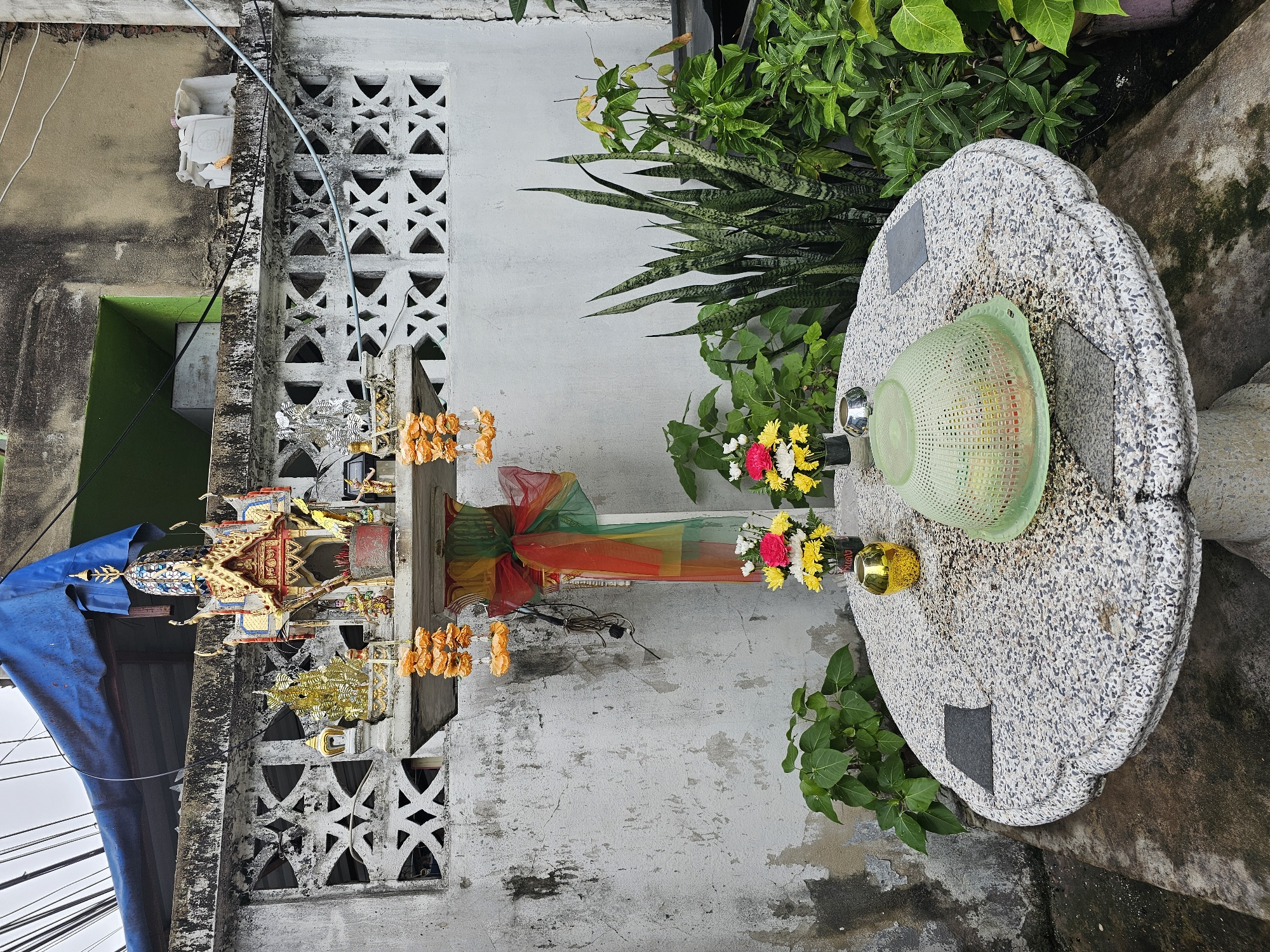 A small, ornate spirit house, common in Thai culture, is displayed on a pedestal against a textured, weathered concrete wall in Bangkok, Thailand. The spirit house is adorned with gold trim, colorful mosaic tiles, and miniature figures, with strings of orange marigold garlands hanging from it. A red, green, and yellow ribbon is tied around the pedestal. In front of the wall, a round, speckled granite table holds two small vases of flowers, a golden ornament, and a green plastic colander filled with various items. Lush green plants, including a snake plant, surround the table. Overhead, electrical wires are visible against a cloudy sky, and a blue tarp is draped over a section of the building.