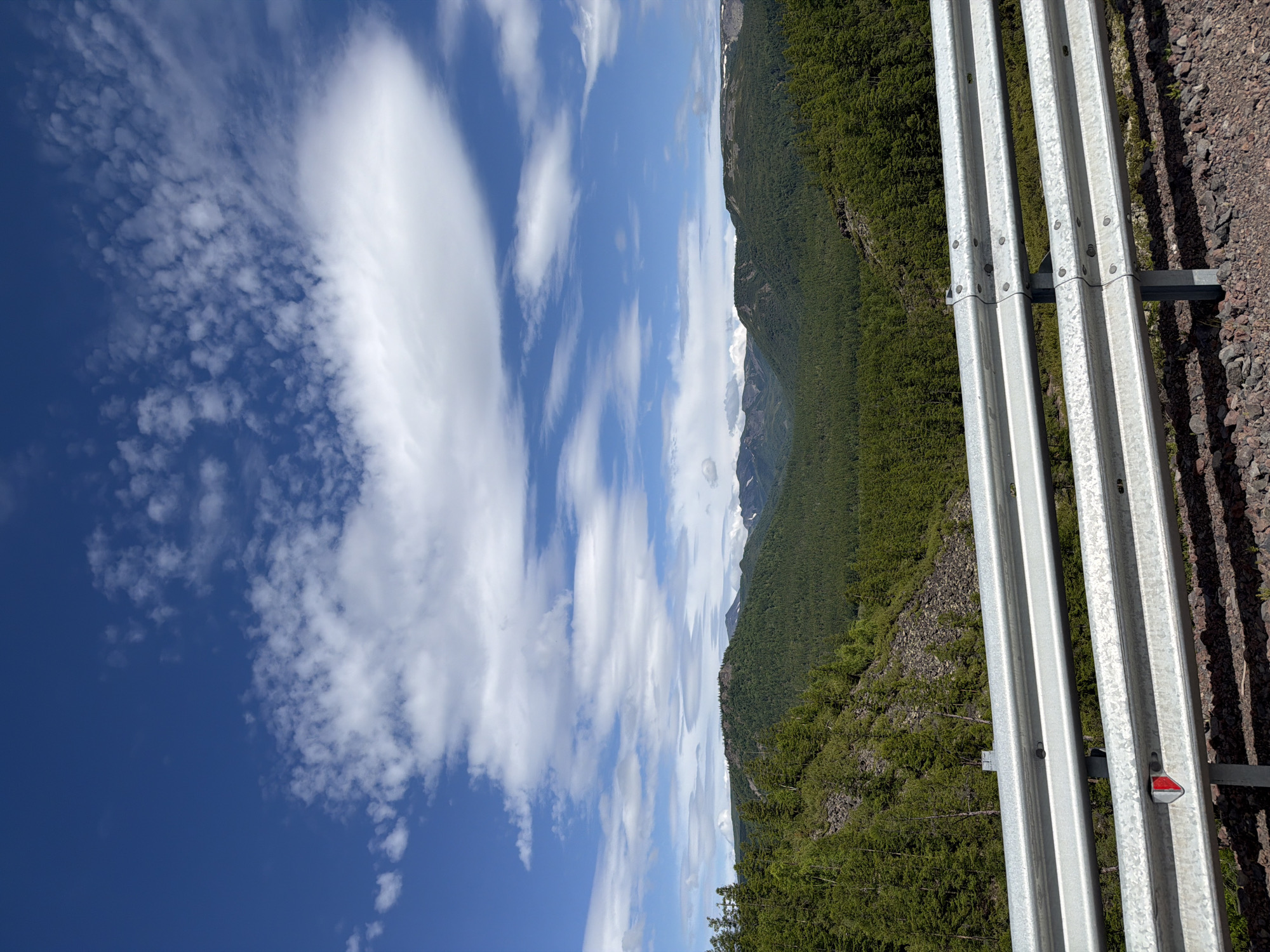 Anavgai, Russia - A scenic vista overlooks a vast expanse of rolling, tree-covered mountains under a partly cloudy blue sky. In the foreground, a silver metal guardrail with three horizontal bars runs across the bottom of the frame, separating the viewer from the mountainous landscape. The guardrail is attached to vertical metal posts, and a small red reflector is visible on the bottom bar. The ground in front of the guardrail consists of dark, gravelly soil. The mountains in the distance are covered in dense green coniferous forest, with some areas of rocky, barren slopes. In the far distance, the peaks of some mountains appear to have patches of snow. The sky is a vibrant blue, dotted with scattered white cumulus clouds, some of which are elongated and wispy.