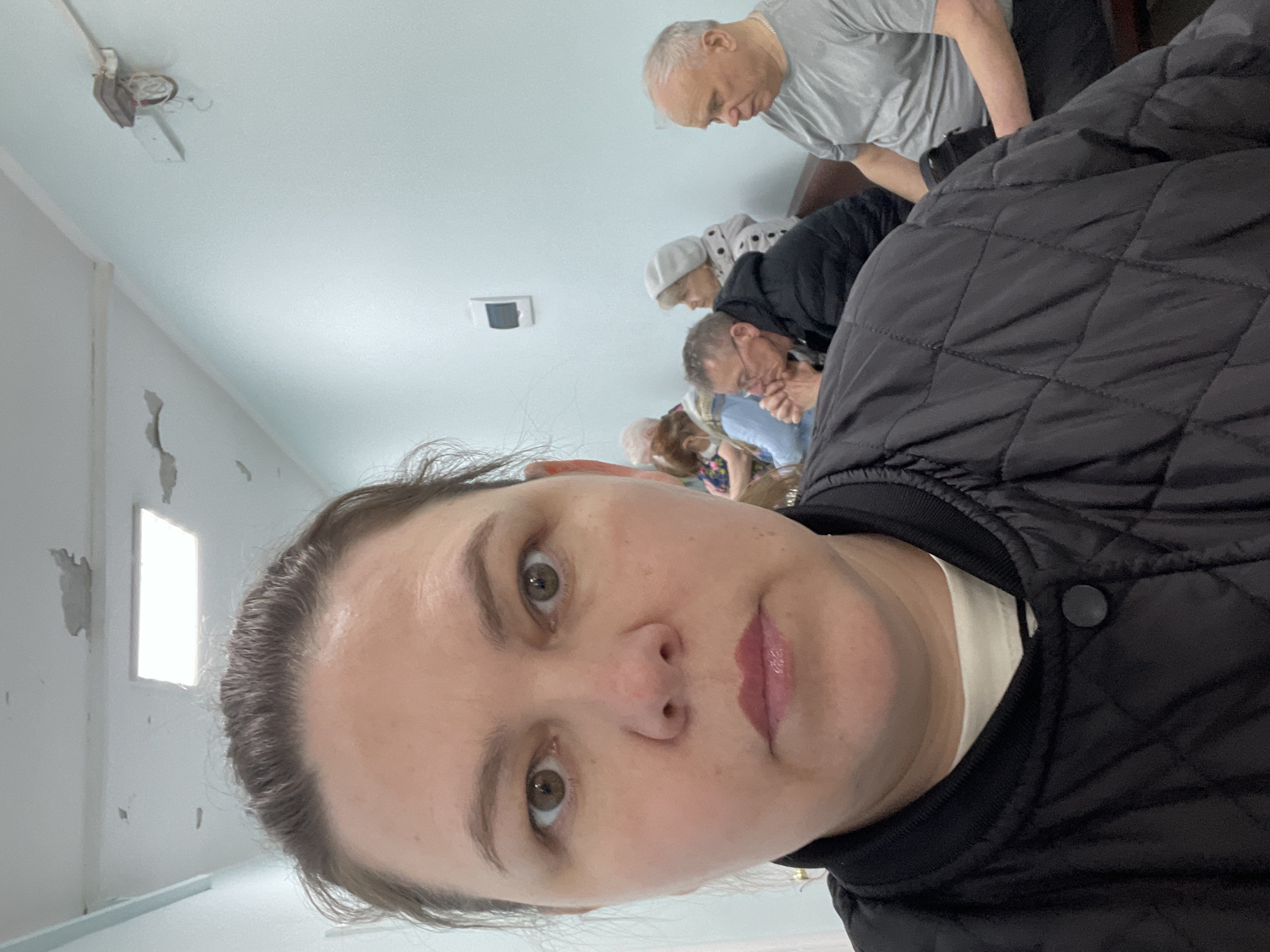 A selfie taken indoors captures a woman's face in the foreground, with several individuals and the interior of a building visible in the background. The woman, with light brown eyes and a neutral expression, wears a black quilted jacket over a white top.
Behind her, multiple people, primarily older adults, are seen leaning forward and looking downwards, indicating their attention is focused on something below the camera's view. A man with short grey hair and a grey t-shirt is visible, alongside an older woman wearing a white head covering.
The setting appears to be a functional indoor space in Noviy, Russia, possibly a hall or communal room. The light-colored walls show some signs of wear, including peeling paint. A small, brightly lit rectangular window is visible on a far wall, contributing to the daytime illumination. The ceiling has a visible, somewhat rudimentary light fixture with exposed wires and a separate rectangular vent. No text is visible in the image. The collective posture of the background figures suggests they are observing an event or participating in an activity requiring their focused attention.