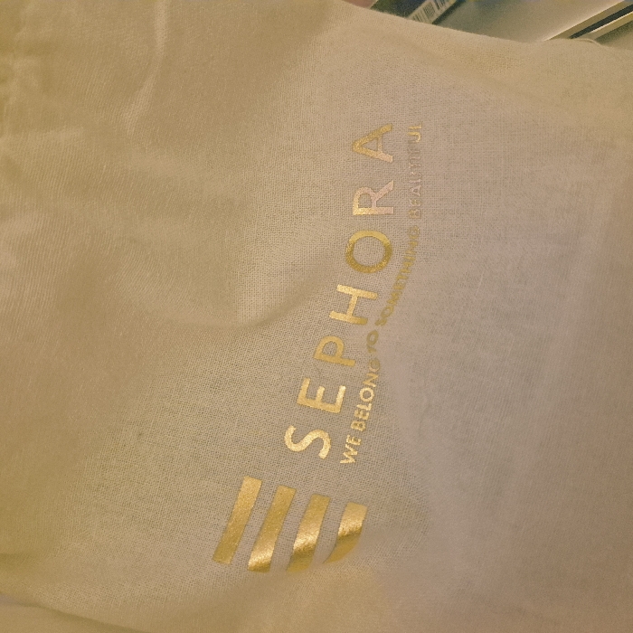 The image displays a light beige fabric bag or pouch with gold metallic branding. The word "SEPHORA" is prominently displayed across the central area in capital letters. Below it, in smaller gold text, reads "WE BELONG TO SOMETHING BEAUTIFUL." To the left of "SEPHORA" is a gold stylized logo consisting of four horizontal bars of varying lengths. The fabric exhibits a fine weave texture. The background is indistinct and offers no specific visual cues to identify a precise location within Strasbourg, France, beyond the presence of a Sephora branded item, a retail chain known to have outlets in the city. No people or specific actions are visible in the scene.