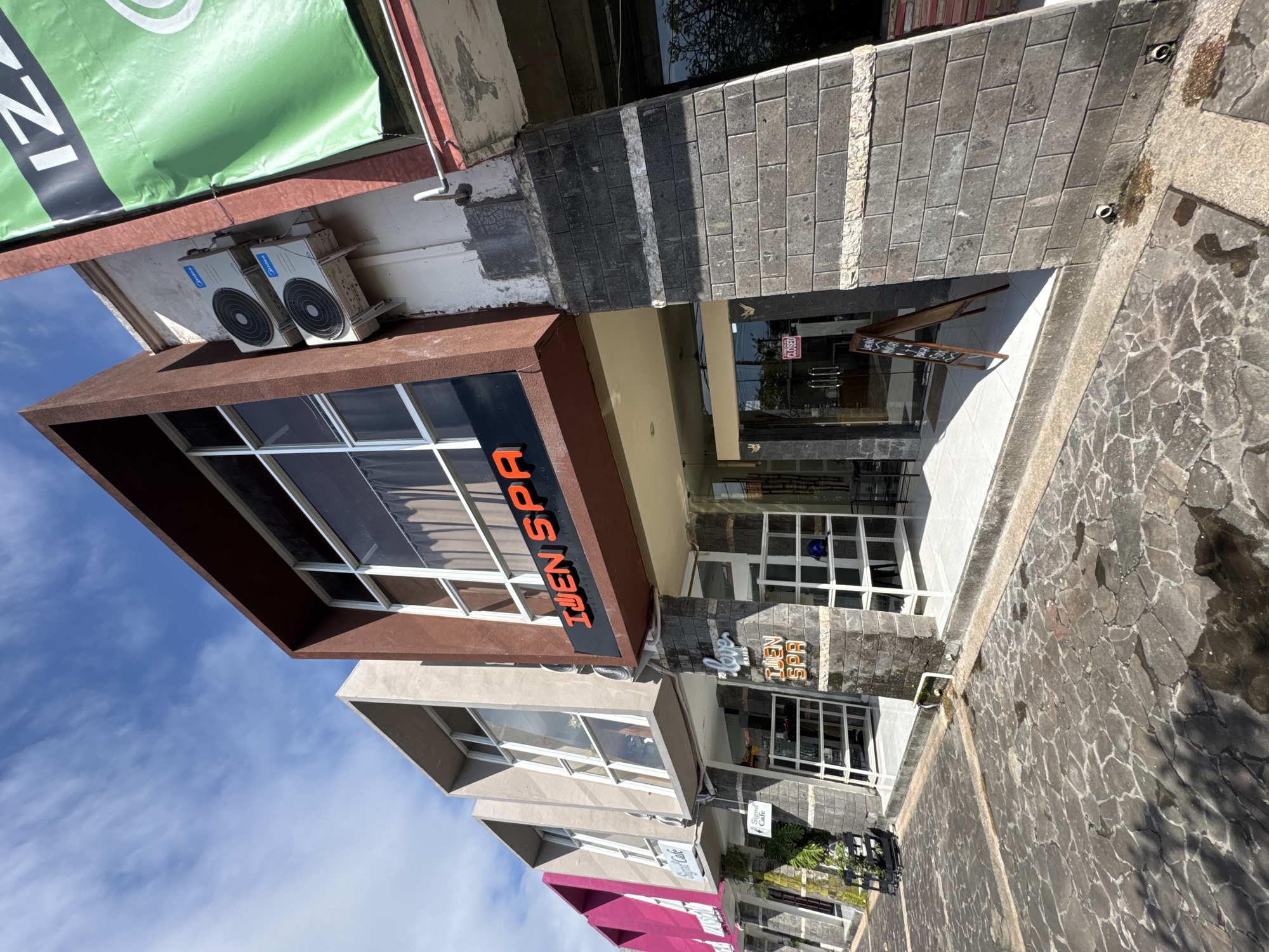 The image depicts a street-level view of a building in Kuta Selatan, Indonesia. The building houses "Ijen SPA," identifiable by signage on the facade. The building has a brown overhang with large windows. Two air conditioning units are attached near the top of the building. A banner is partially visible on top of the building. A sidewalk and paved street are present in front of the building, and another business called "Sigma Cafe" is also visible on the street. There are no people visible in the image.