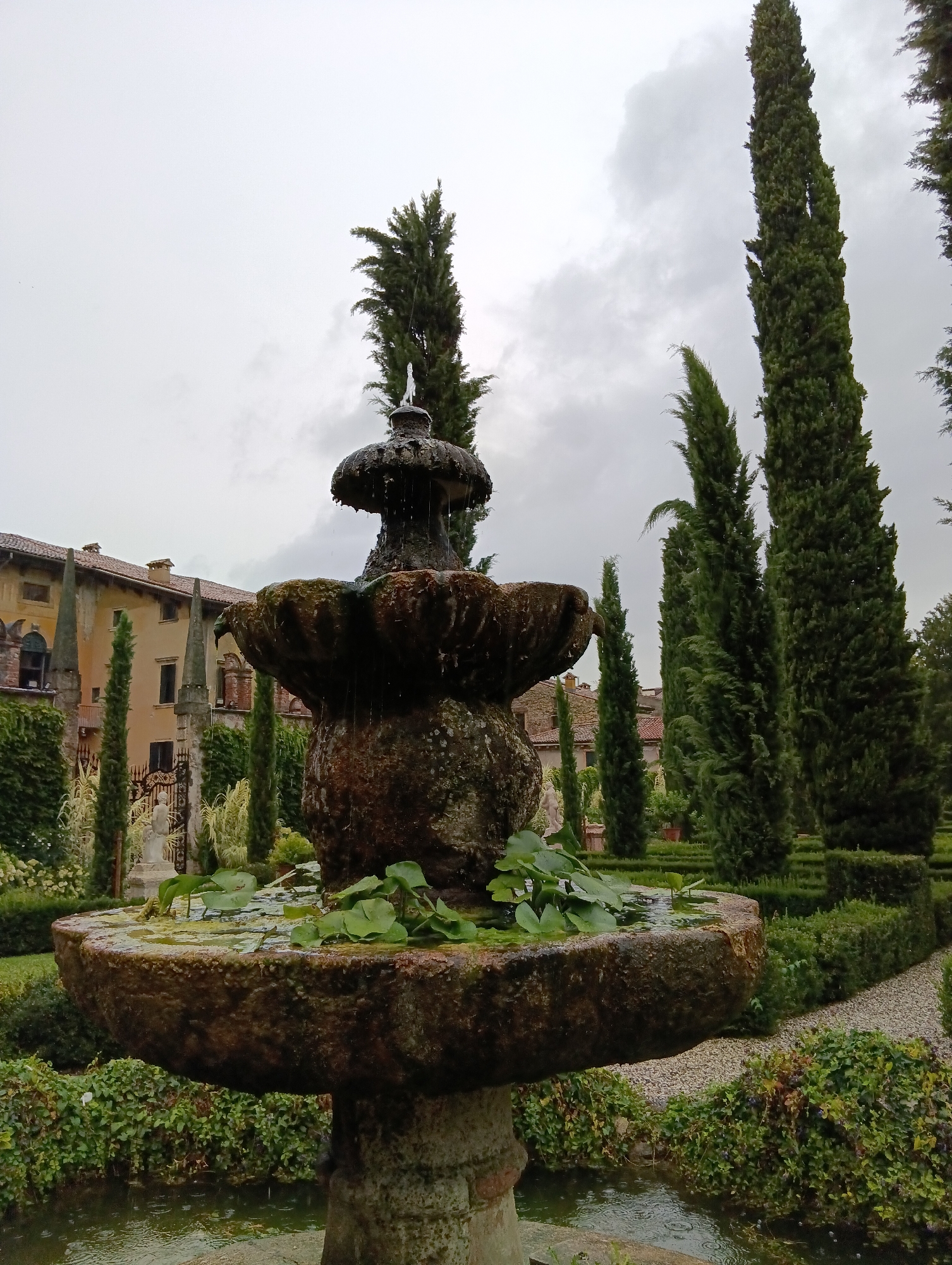 The image depicts an old stone fountain in a garden. The fountain is the central subject, with water flowing from its upper tiers into the basin filled with lily pads. Surrounding the fountain are lush green bushes and manicured hedges, forming a formal garden setting. In the background, a pale yellow building with red tiled roofs and arched windows is visible, suggesting a historical estate or villa. Tall, slender cypress trees frame the scene, reaching towards a cloudy sky. The overall atmosphere is serene and somewhat melancholic, possibly due to the overcast weather. This scene is characteristic of the gardens found at Villa Arvedi, a historical villa located in the Lessinia hills on the outskirts of Verona, Italy, known for its extensive botanical gardens and fountains. No people are present in the image, and there are no discernible actions or interactions taking place. The focus is entirely on the architecture of the fountain and the surrounding landscape.